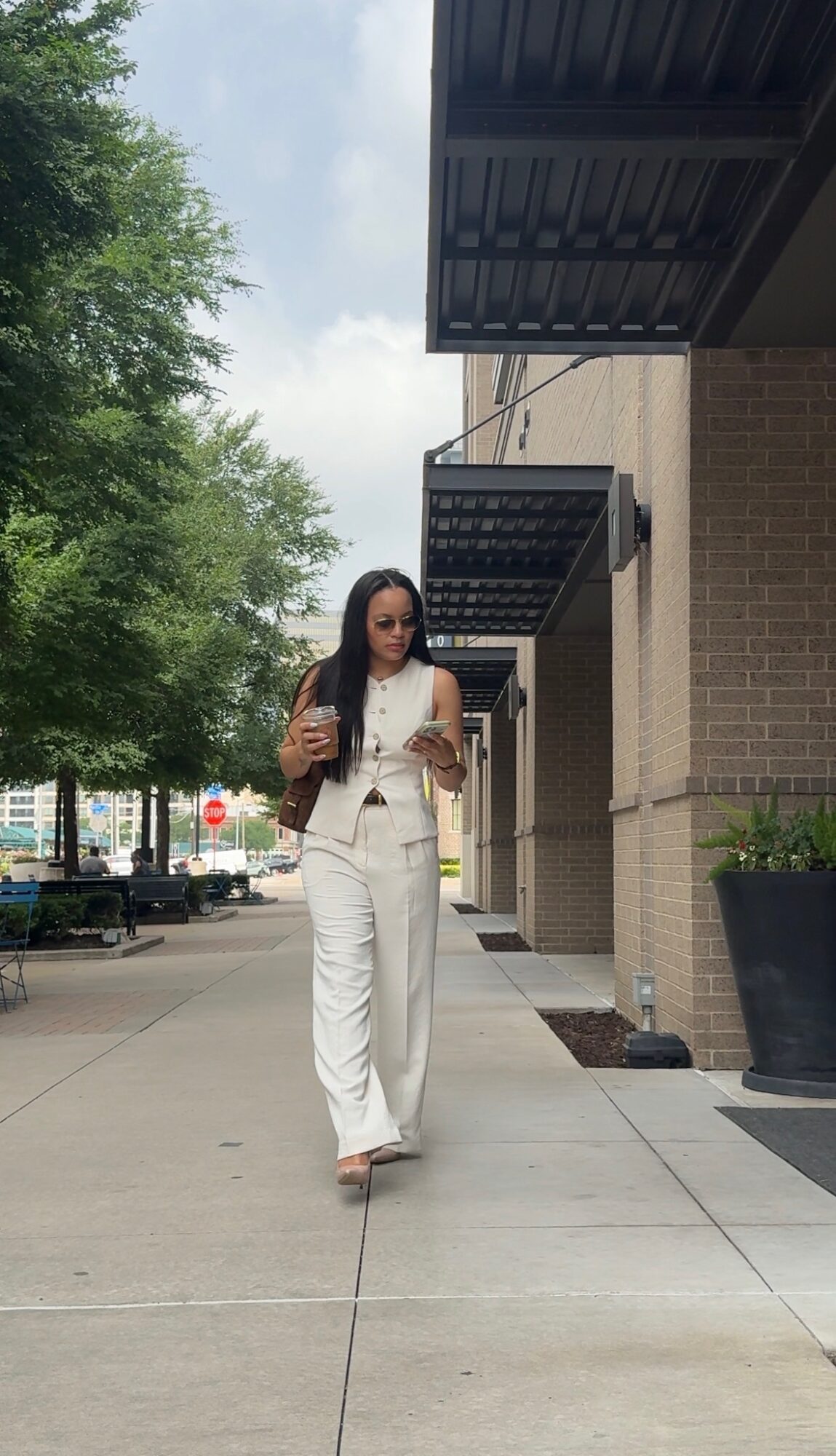 Woman walking on sidewalk in front of building with trees and parked cars in background.