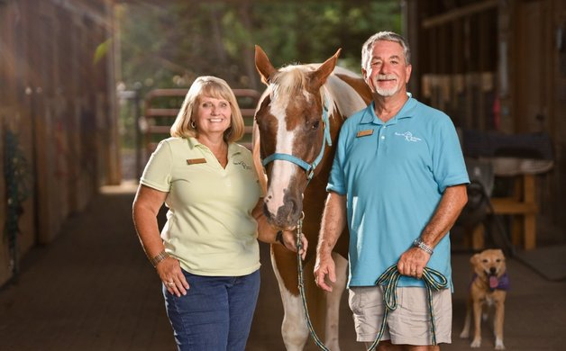 Two people, a woman and a man, stand with a horse inside a barn, with a small dog in the background.