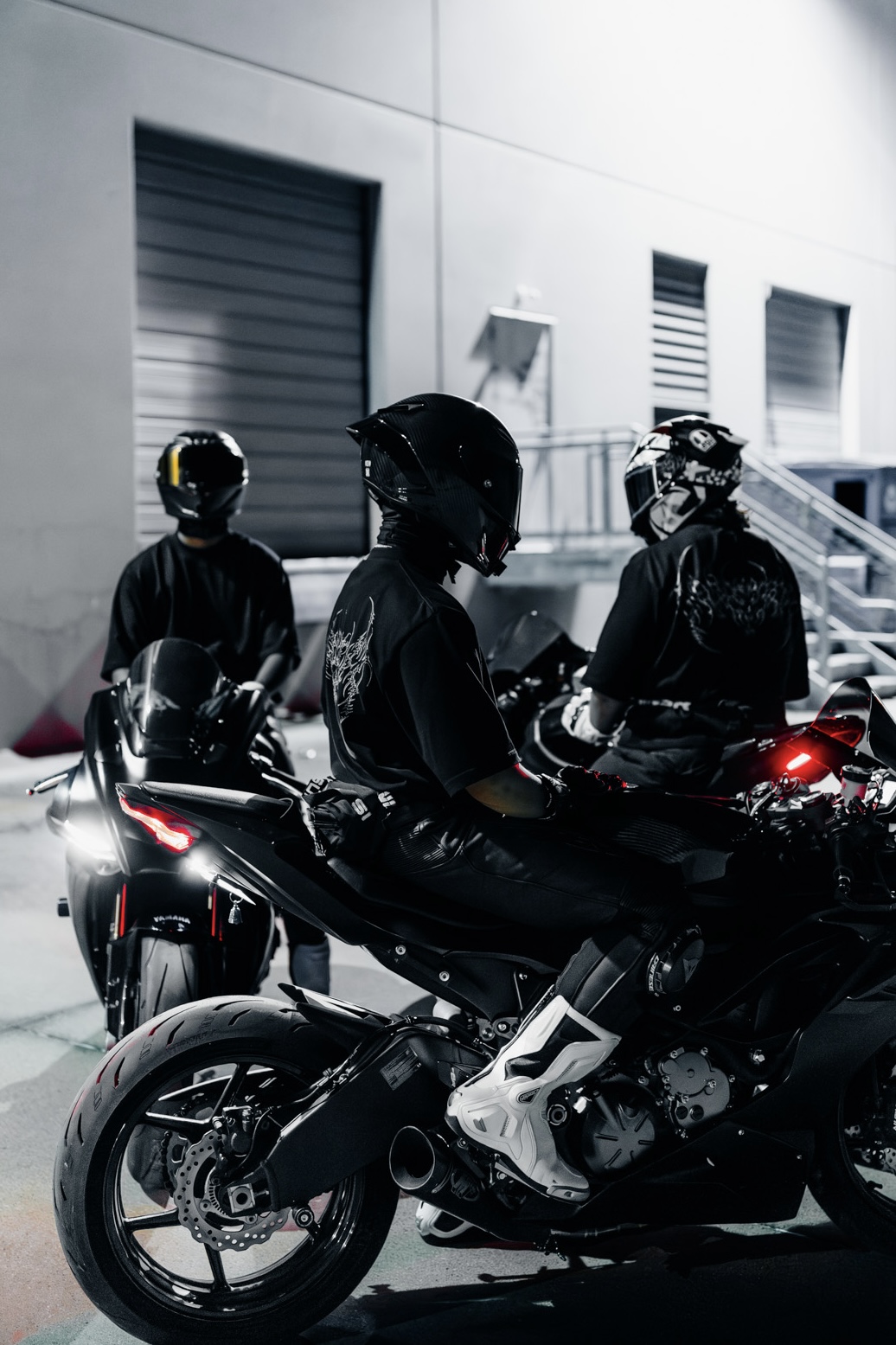 Three motorcyclists wearing helmets and dark clothing sit on motorcycles outside a building with closed shutters.