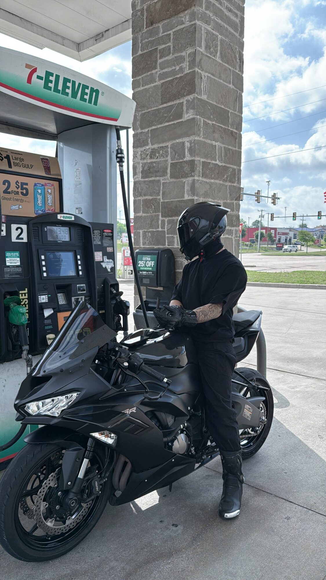 Person in black gear refueling a motorcycle at a 7-Eleven gas station, with a brick pillar and blue sky in background.