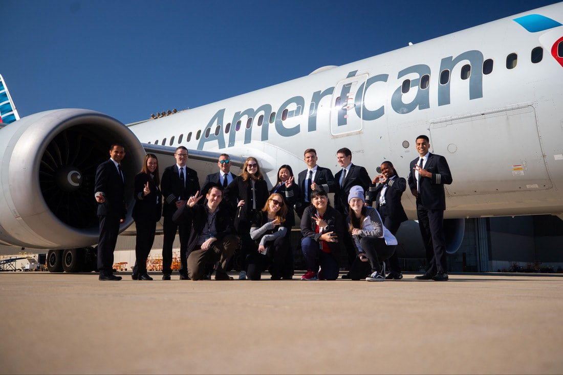 Group of people in business attire posing in front of an airplane with 'American' written on it, outdoors under a clear sky.