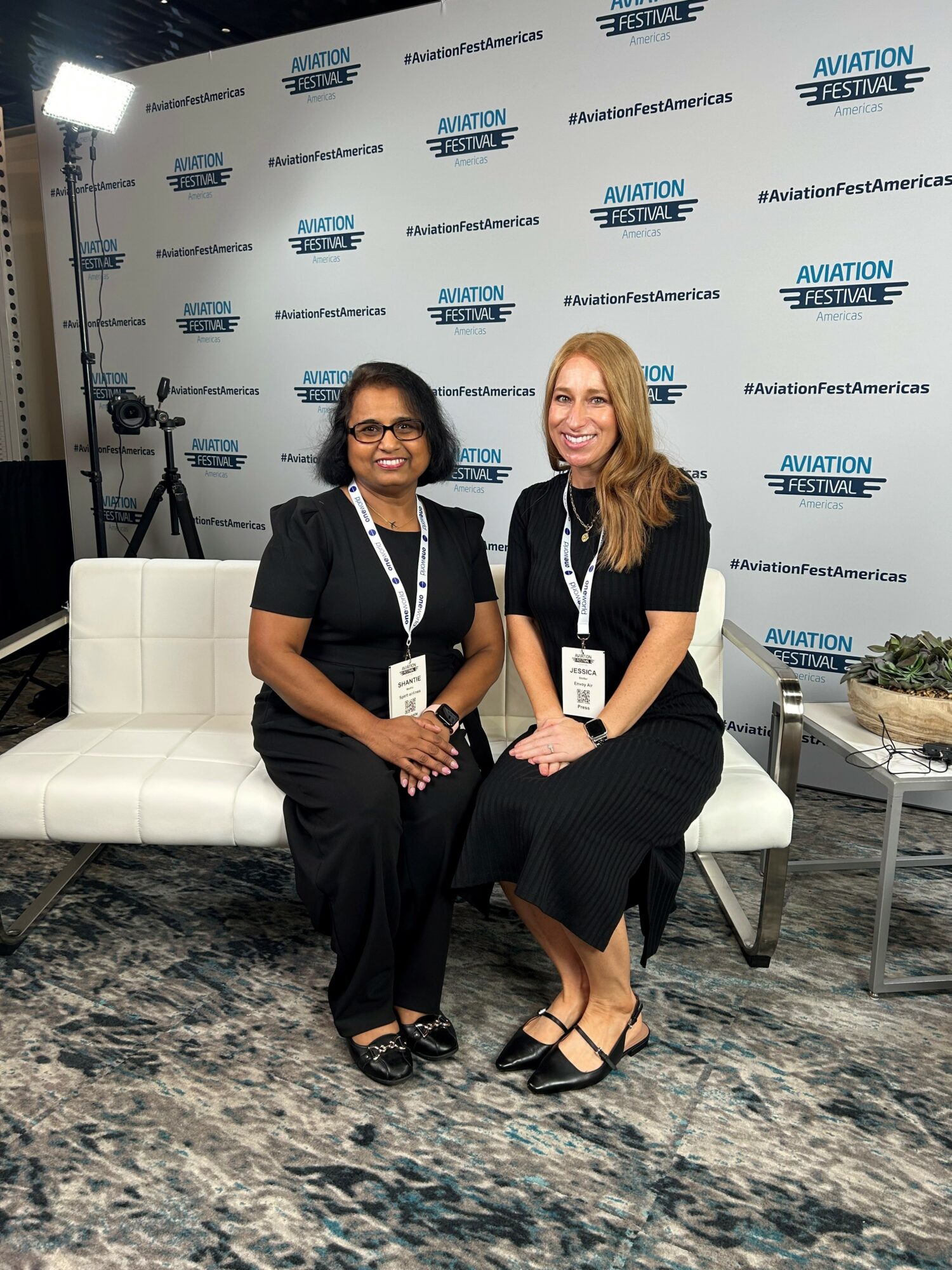 Two women sitting on a white bench at an event, with a backdrop displaying logos and text, smiling at the camera.