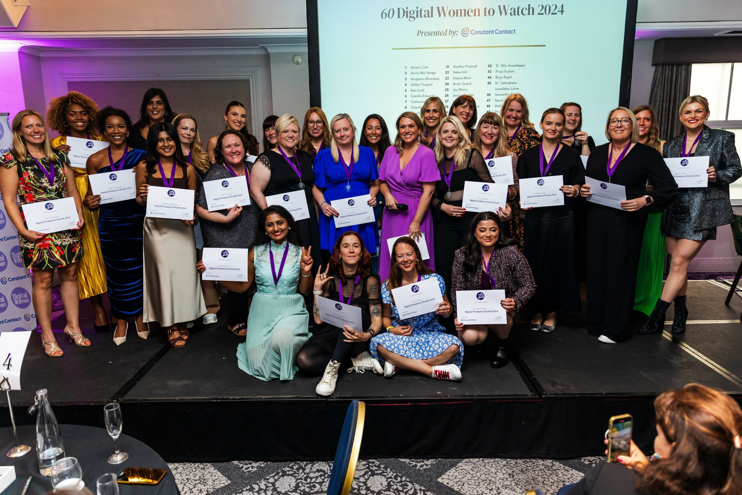 Group of women and girls on stage holding certificates, some wearing medals, in a formal event setting.