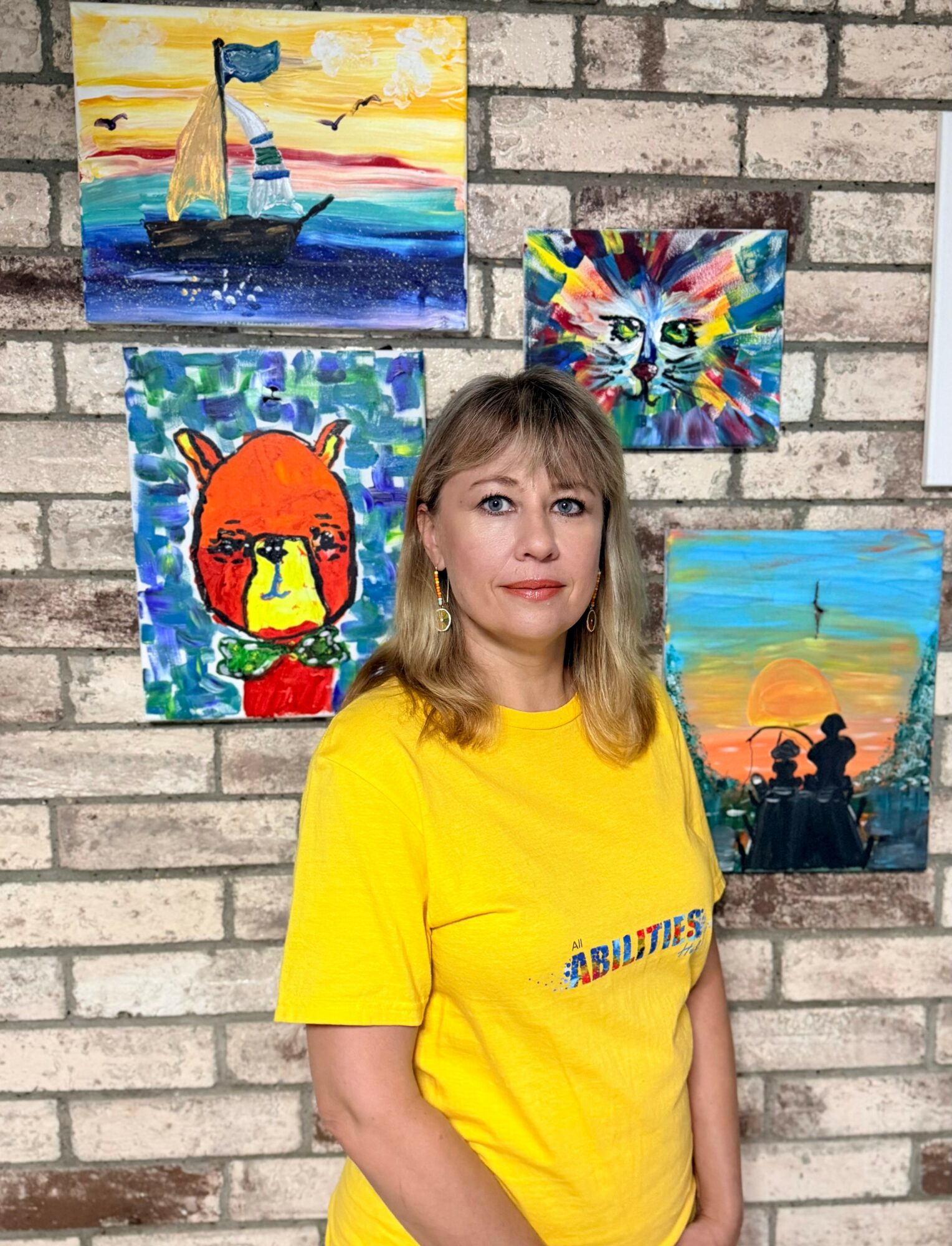 Woman with shoulder-length hair wearing a yellow T-shirt standing in front of a brick wall with colorful paintings.