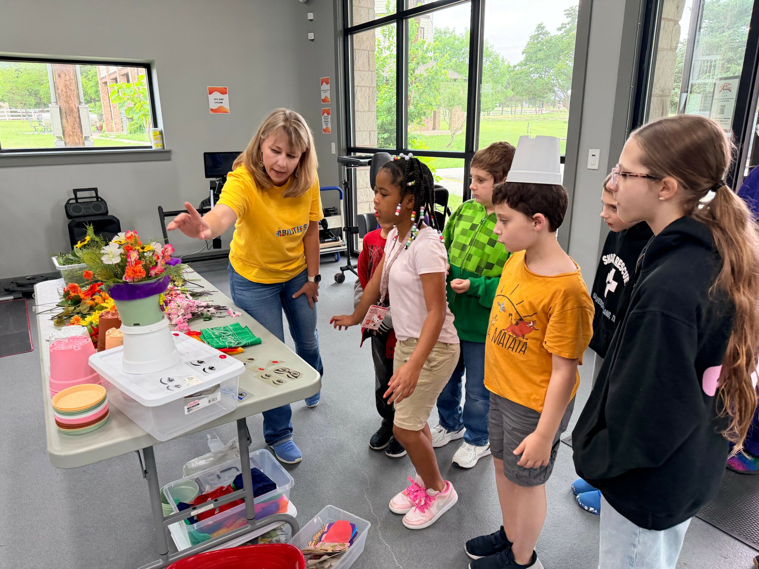 A woman in a yellow shirt demonstrates craft activities to children in a classroom with large windows and plants.