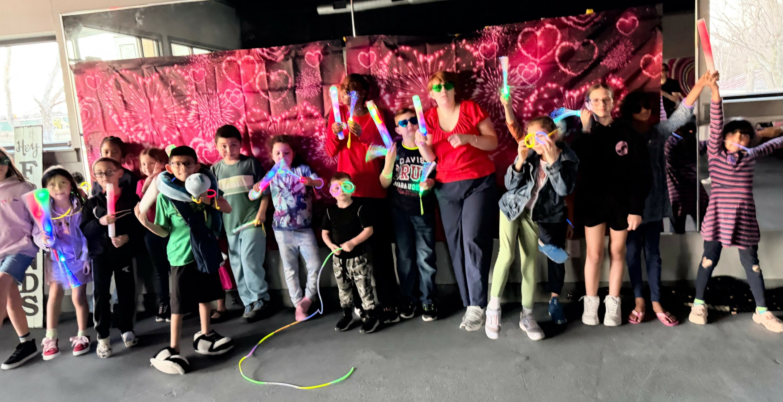 Group of children and adults standing together in front of a pink heart-themed backdrop, some raising hands, in an indoor setting.