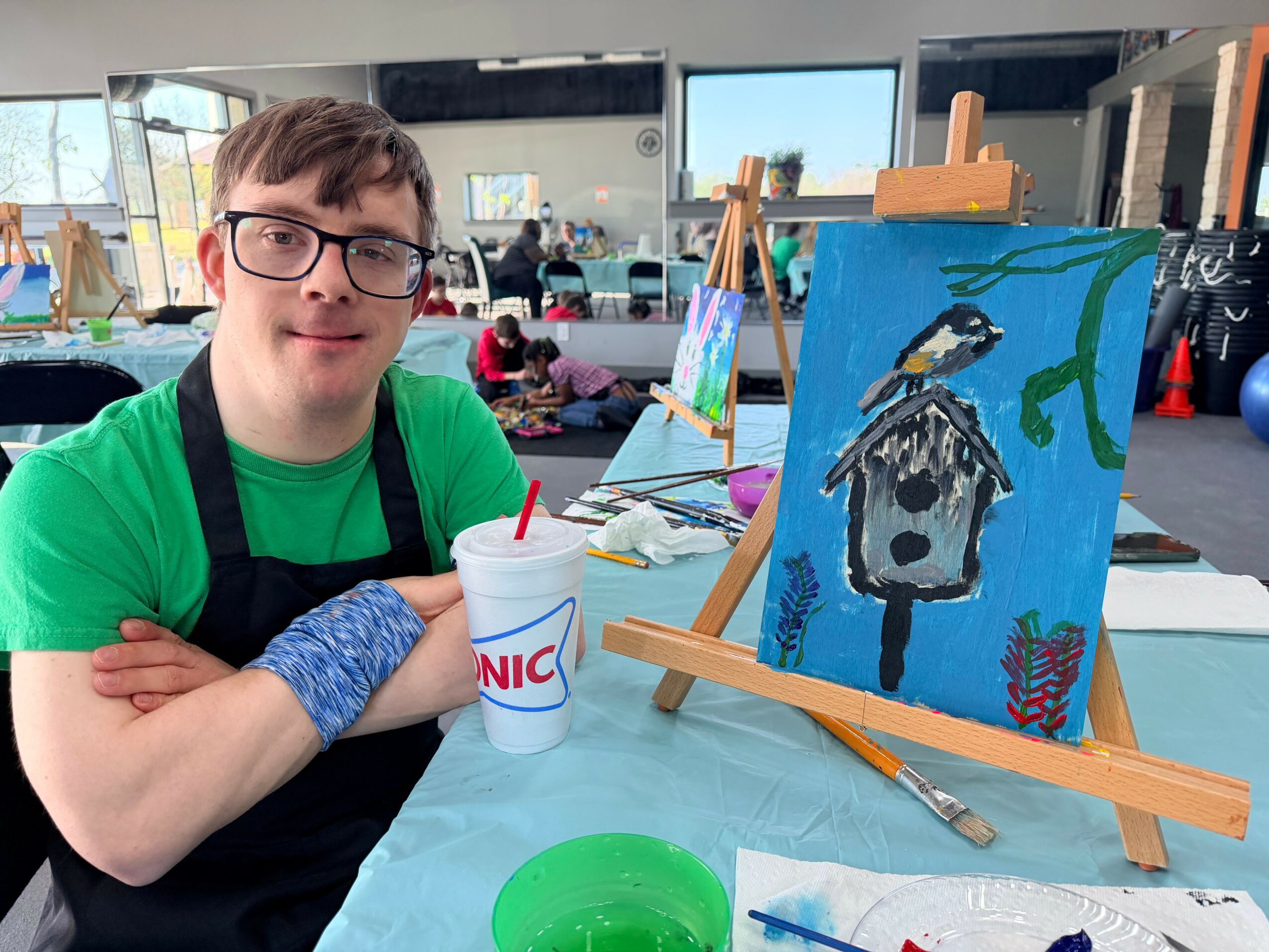 Young man with glasses and a green shirt sits at a table with a painting of a birdhouse on an easel, in a bright indoor space.