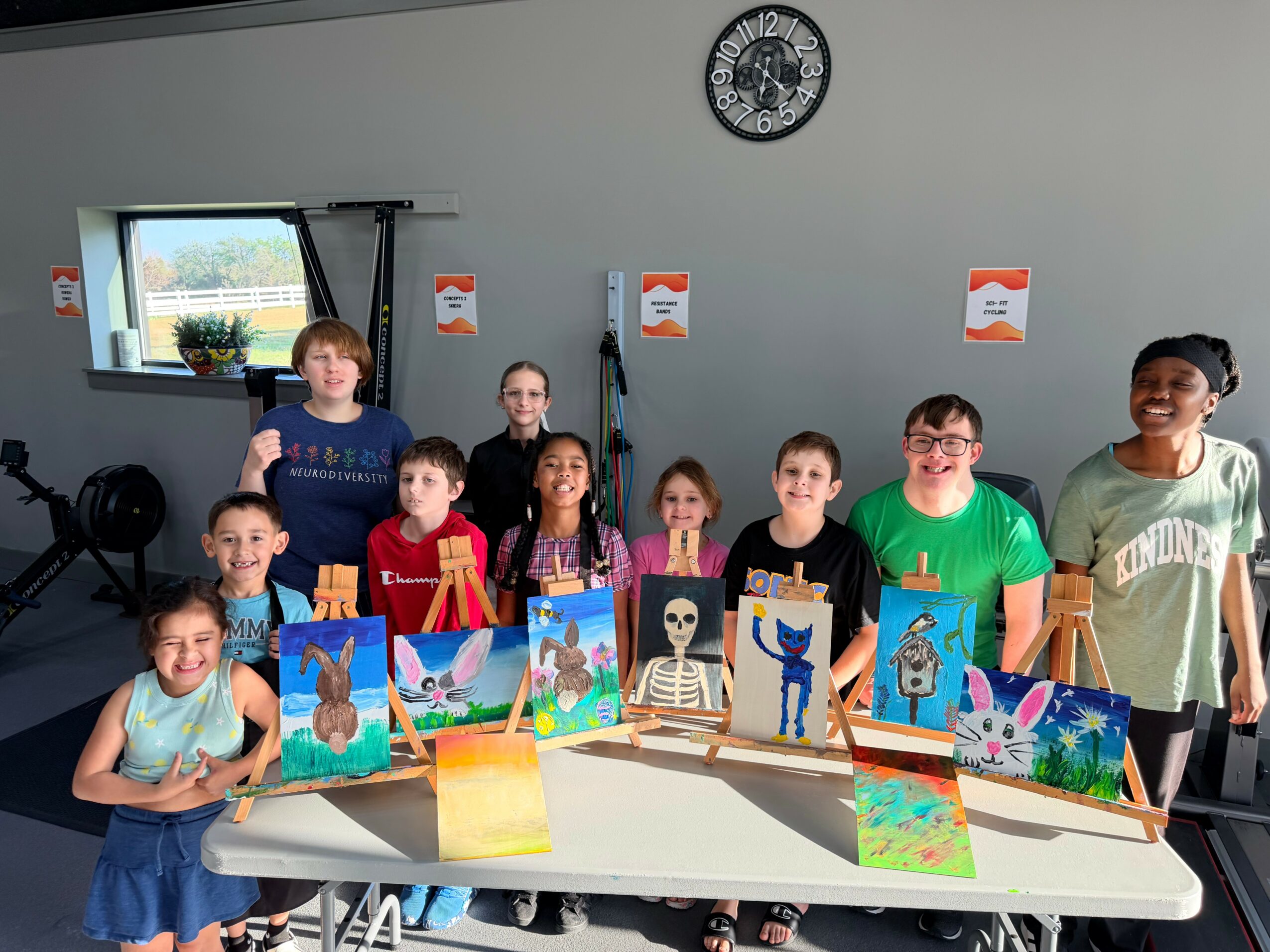 Group of children standing behind a table with colorful paintings, in a room with a clock and window.