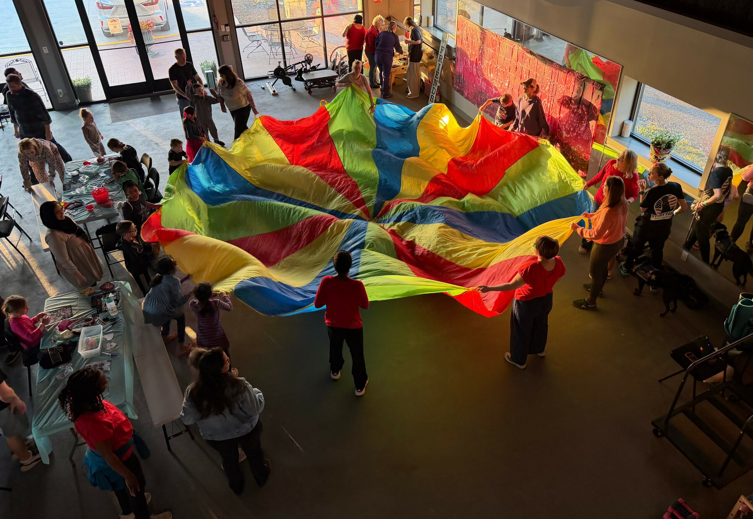 People gather around a large colorful parachute indoors, children and adults holding its edges, with windows and a view outside.