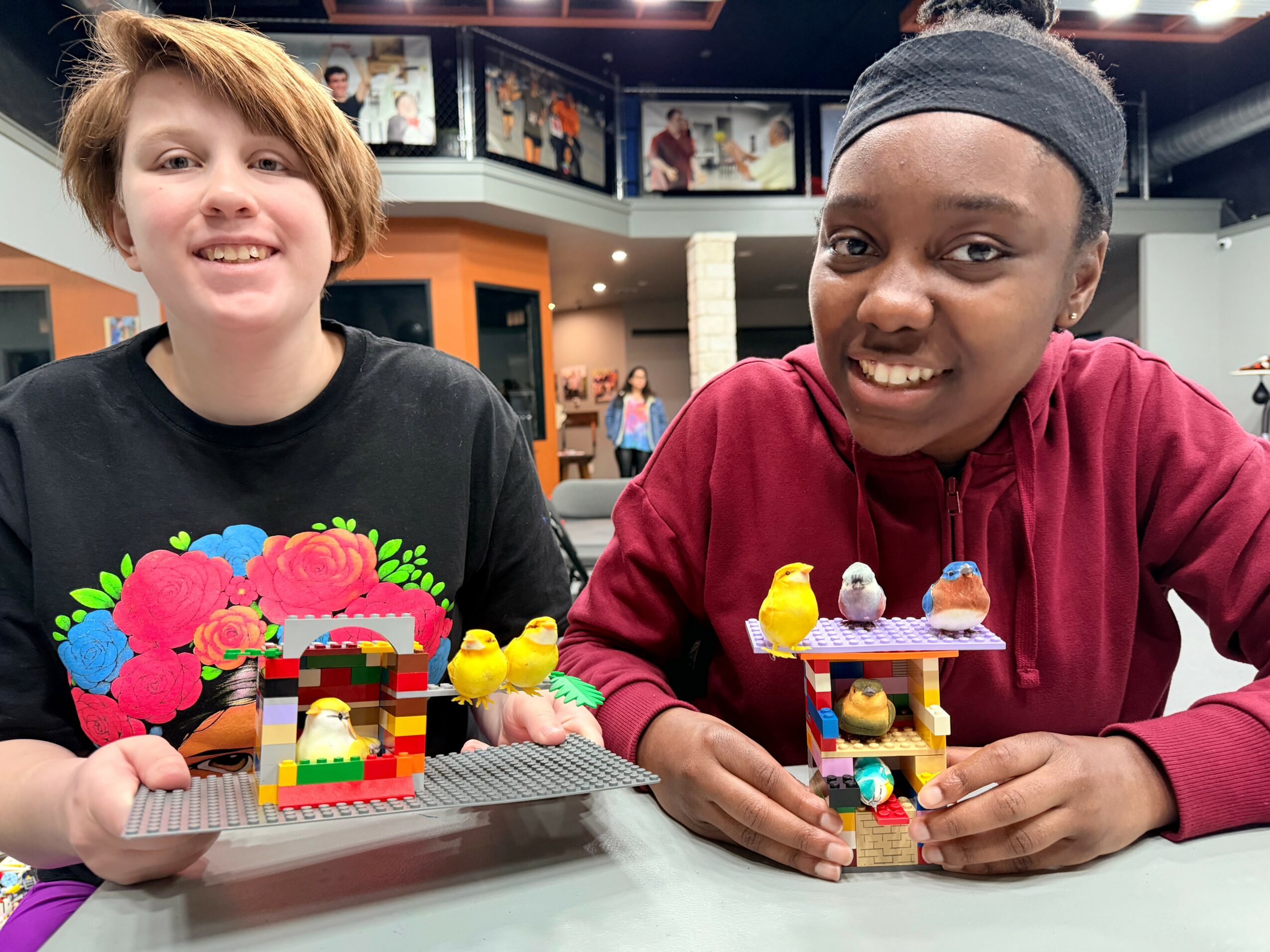 Two children smiling and sitting at a table with colorful LEGO creations in a room with photos on the wall.