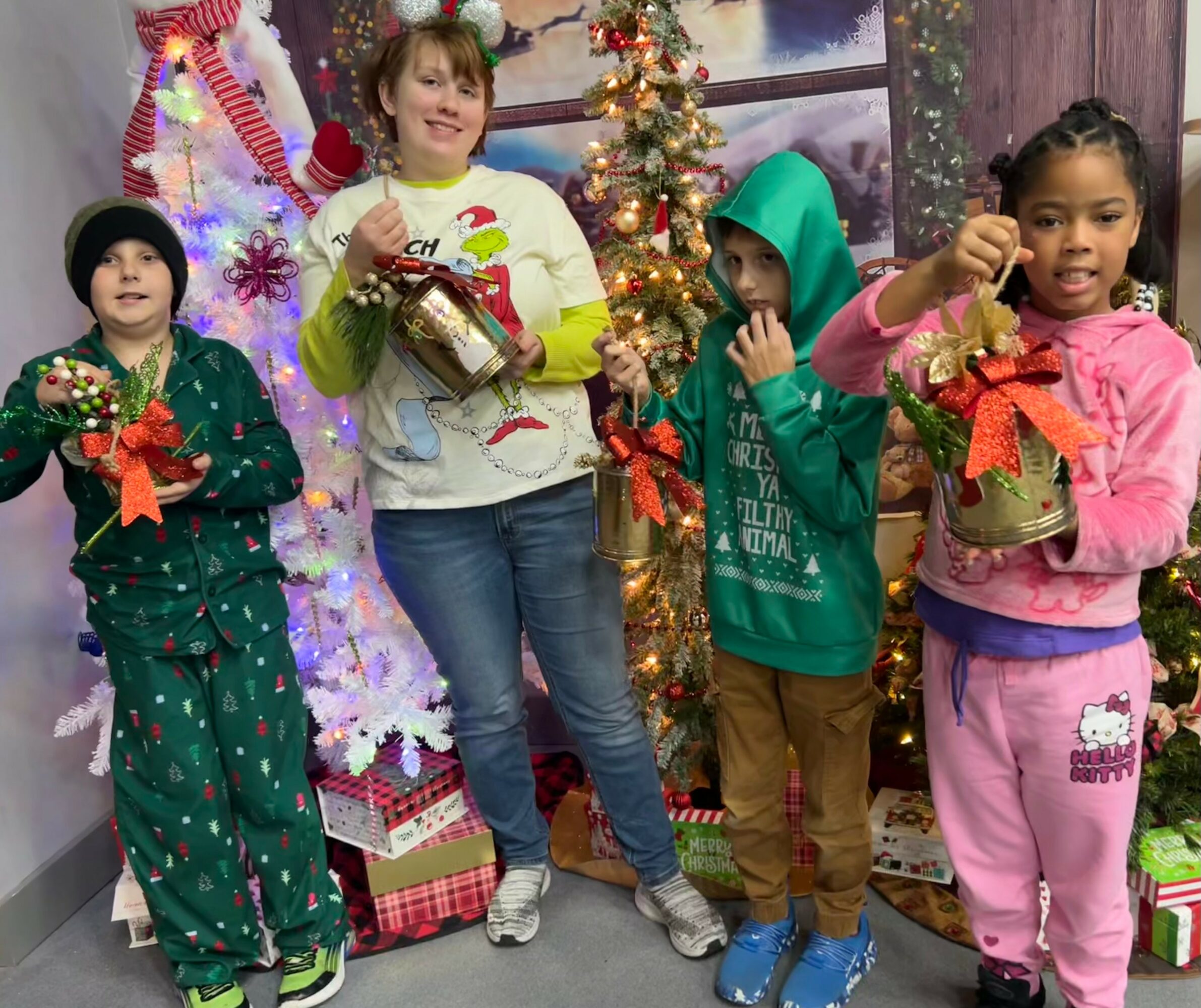 Four children stand in front of a decorated Christmas tree, holding festive jars and gifts, smiling. Christmas decorations are visible.