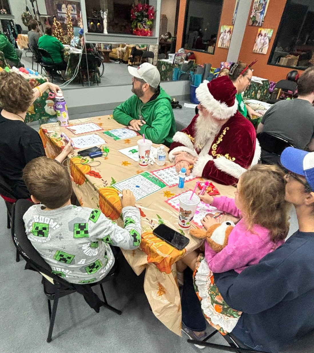 People gathered around a table with holiday decorations, including a person dressed as Santa Claus, children, and adults, in a festive indoor setting.