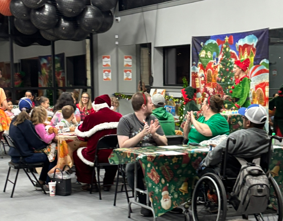 People gather around tables with Christmas decorations, including a person dressed as Santa, in a festive room with holiday artwork.