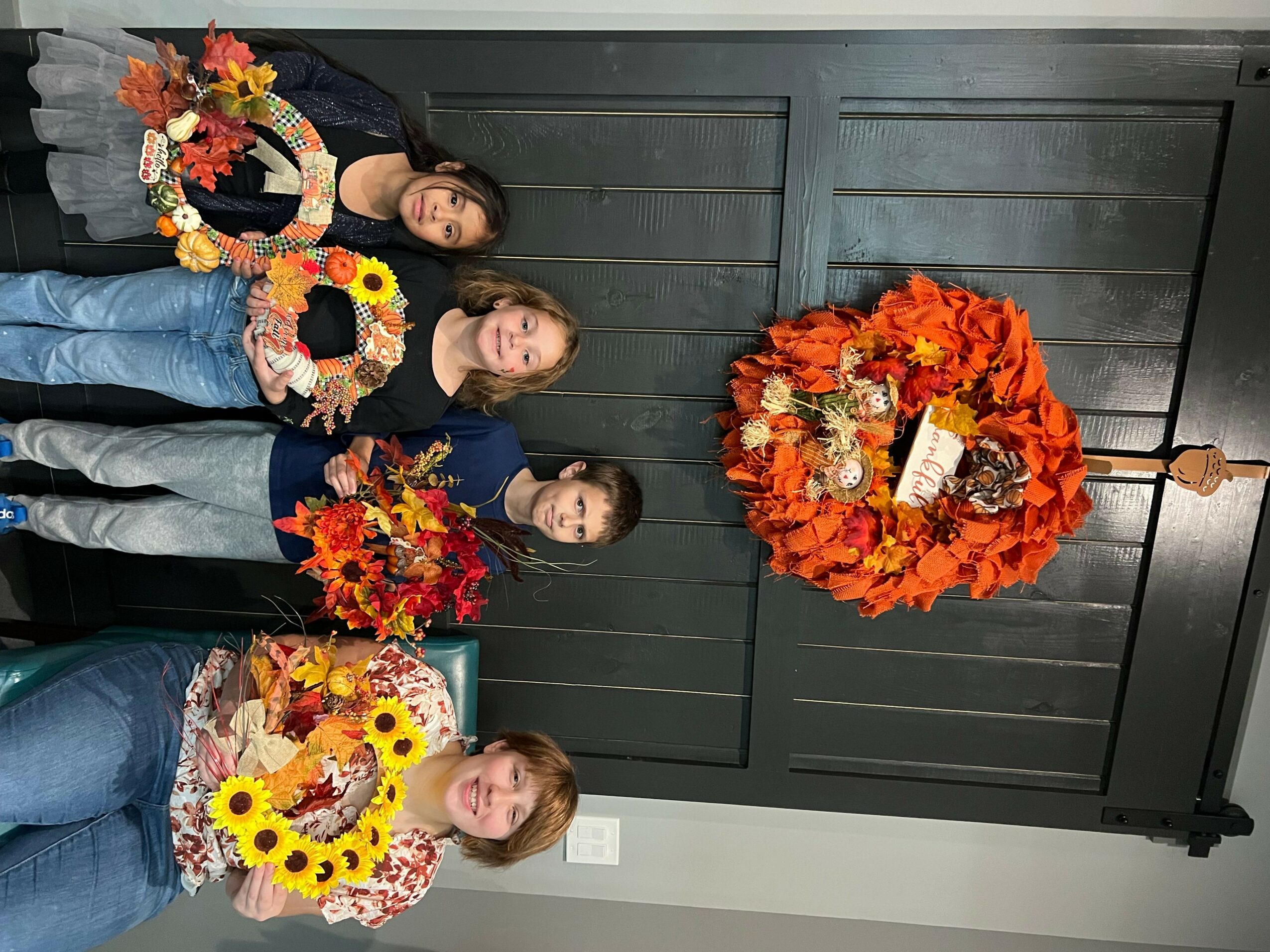 Four people holding floral wreaths and a bouquet, standing in front of a dark wooden door with a large autumn wreath.