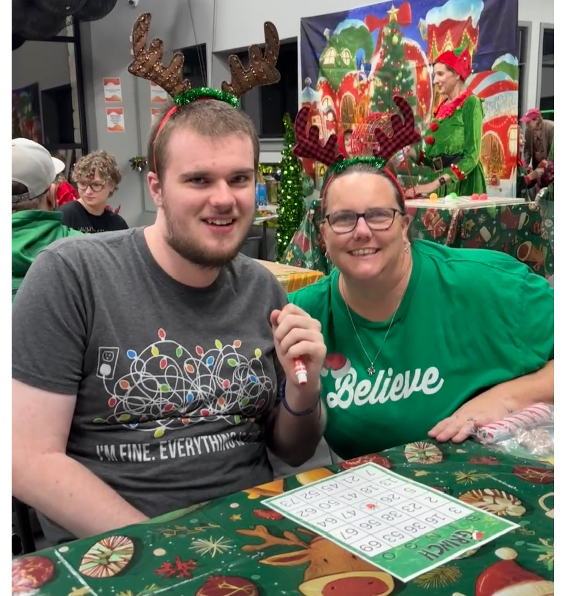 Two people wearing reindeer antler headbands sit at a table with Christmas decorations, smiling, with a festive background behind them.