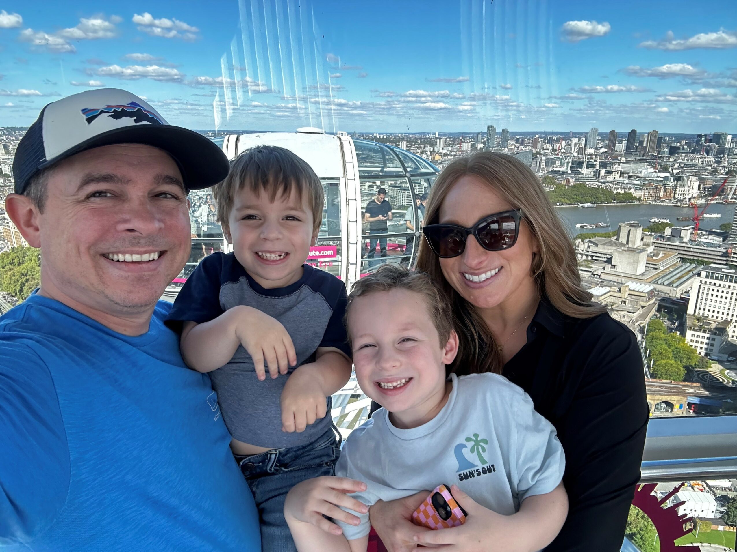 Family of four smiling on a Ferris wheel with cityscape background, two children and two adults, sunny day.