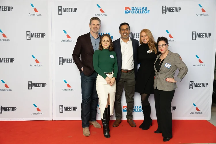 Five people standing on a red carpet in front of a backdrop with logos, smiling for the photo.