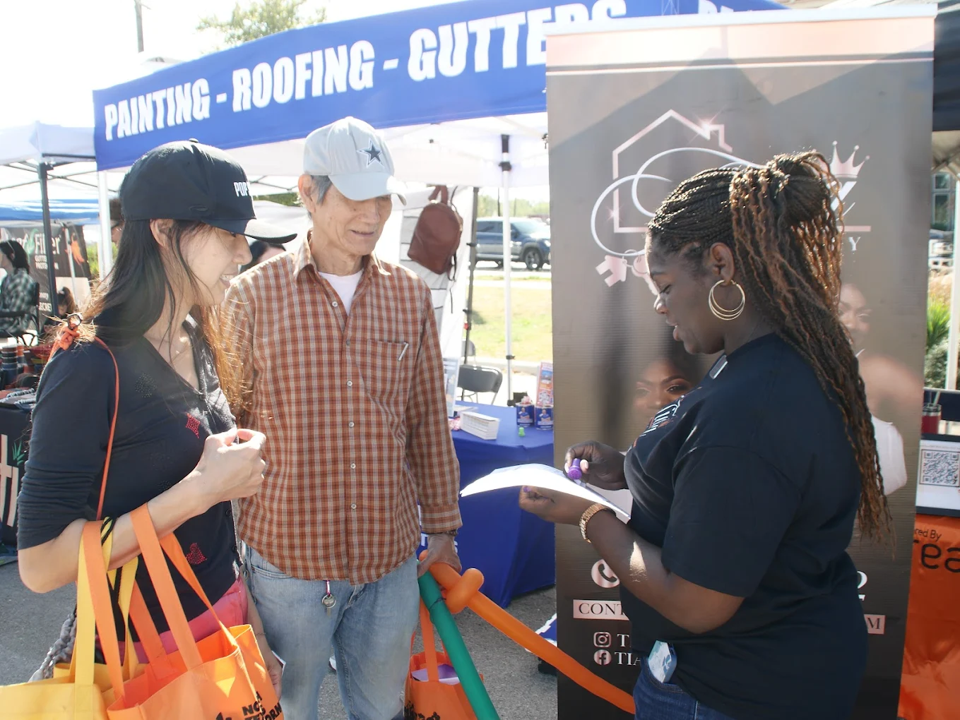 Three people standing outdoors near a booth with a blue sign, two women and one man, engaged in conversation.