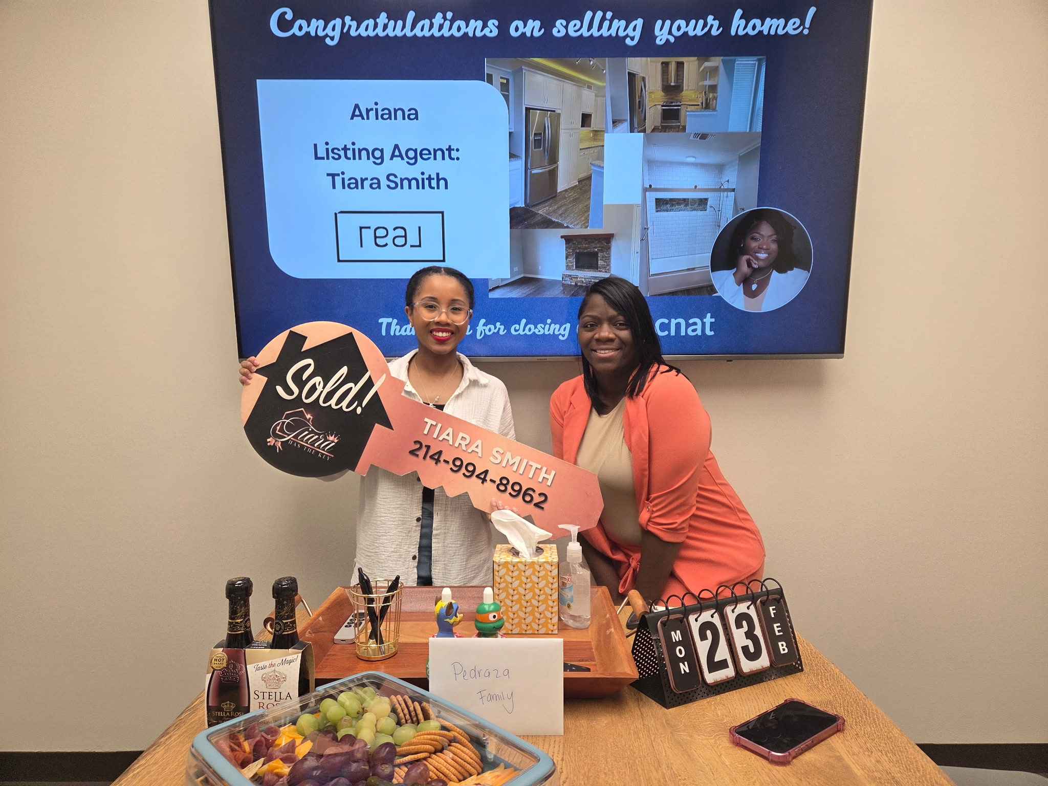 Two women stand behind a table with a large check and various items, in front of a screen displaying a home sale announcement.