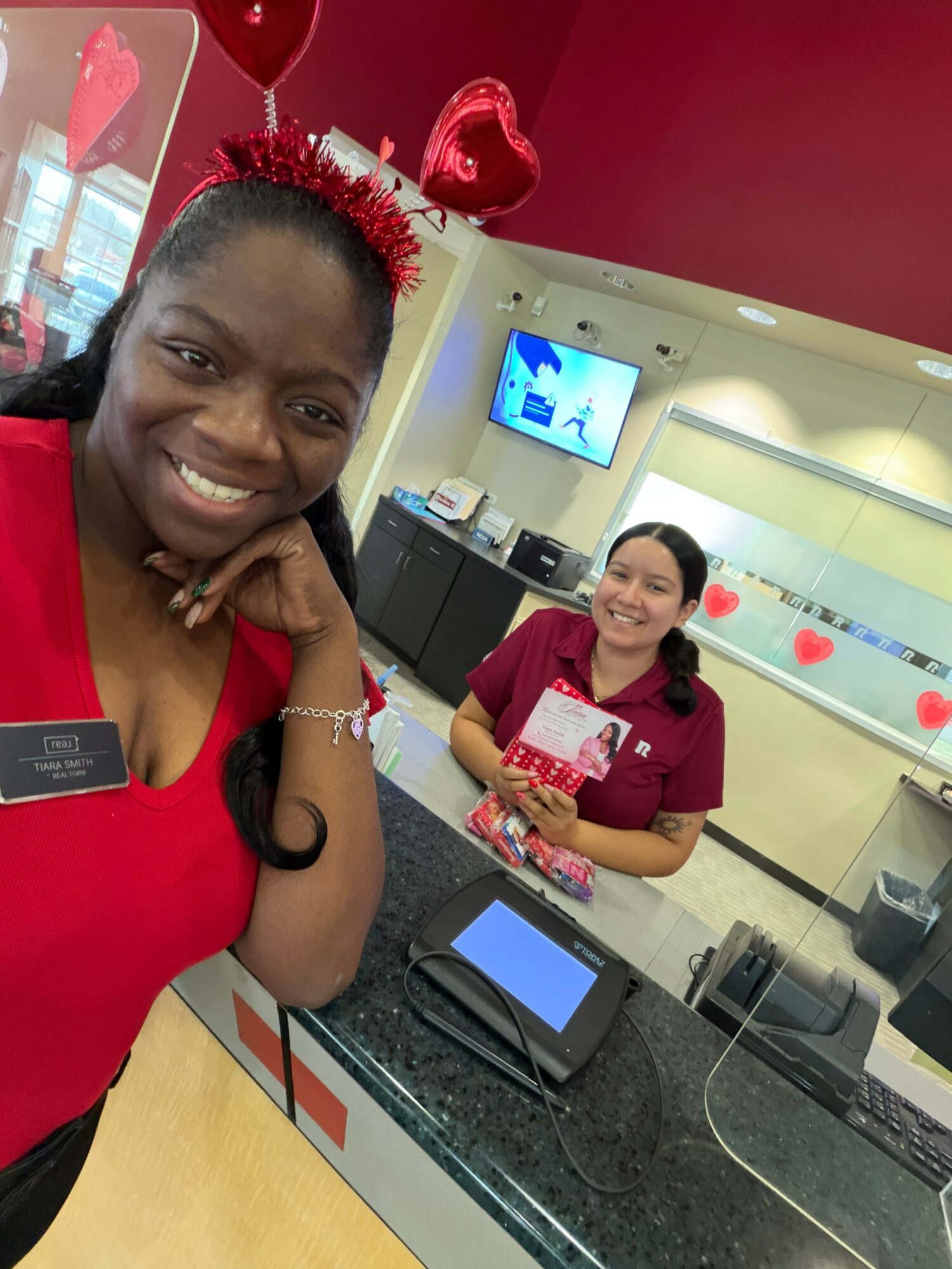 Two women in red uniforms smiling at a counter with Valentine's Day decorations in the background.