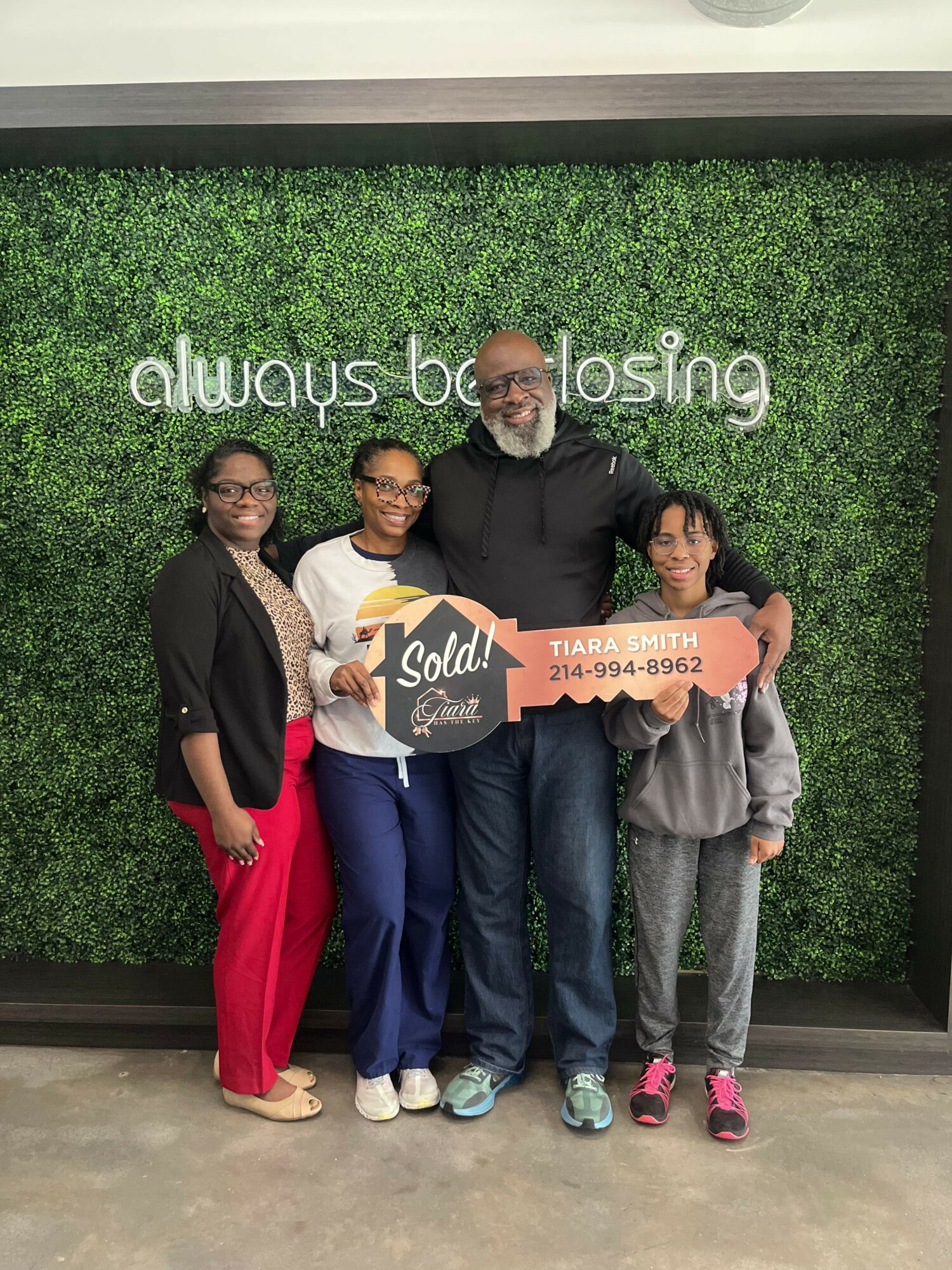 Four people standing in front of a green wall with white text, holding a sign that says 'Soda' and 'Always Losing'.
