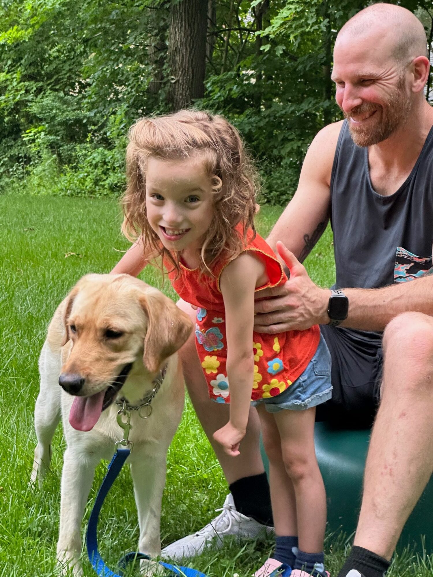 Man, girl, and dog outdoors on grass with trees in background, man holding the girl and dog on leash.