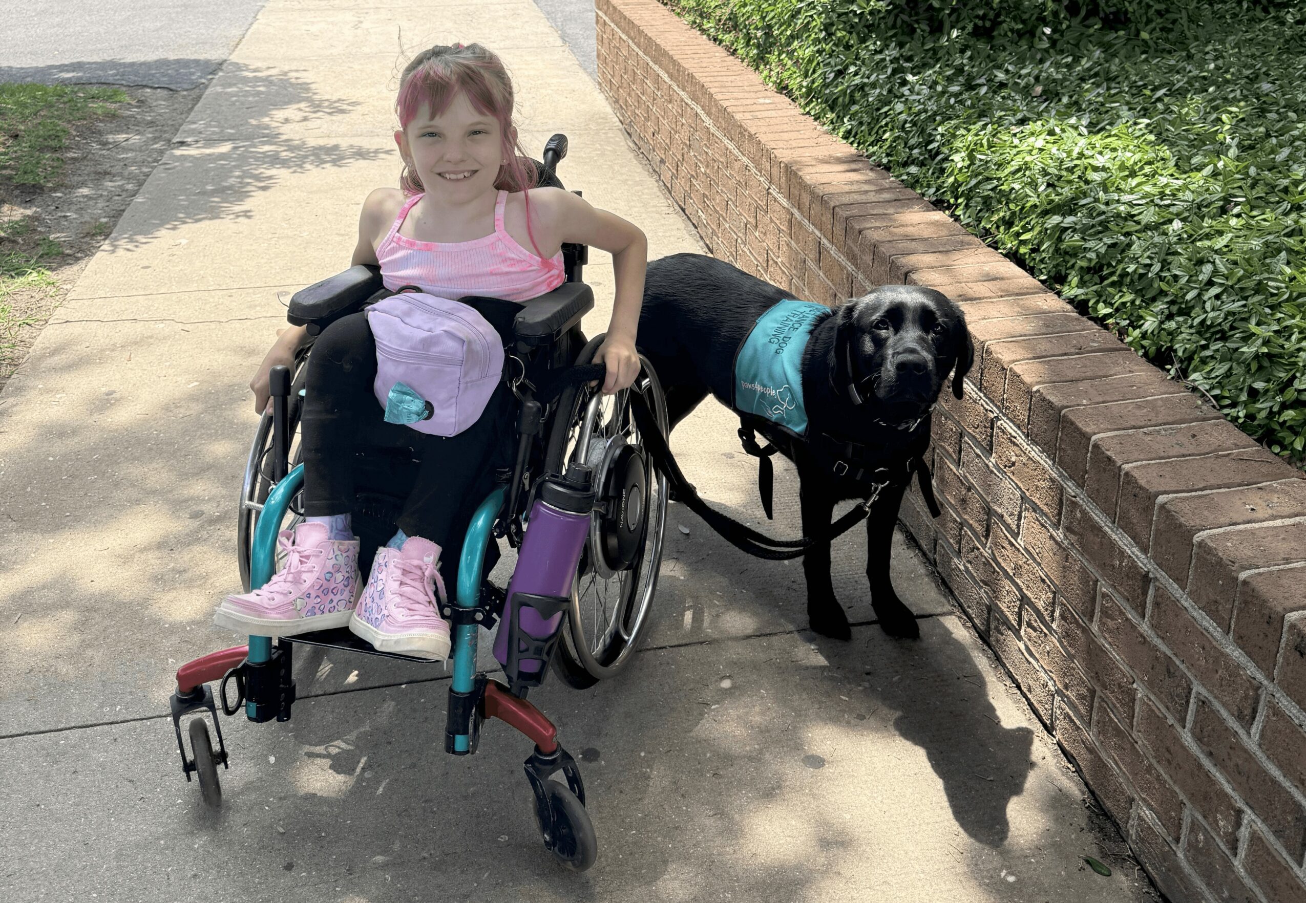 Young girl in a wheelchair smiling outdoors with a black dog beside her, on a sidewalk next to a brick wall.