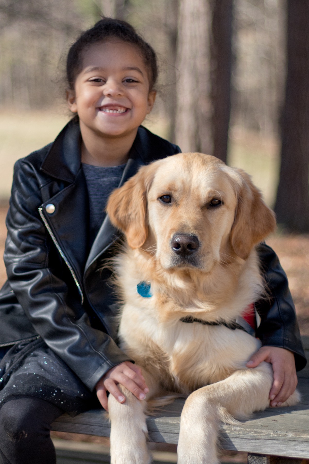 Child smiling with a golden retriever dog outdoors, both sitting on a wooden surface, trees in background.