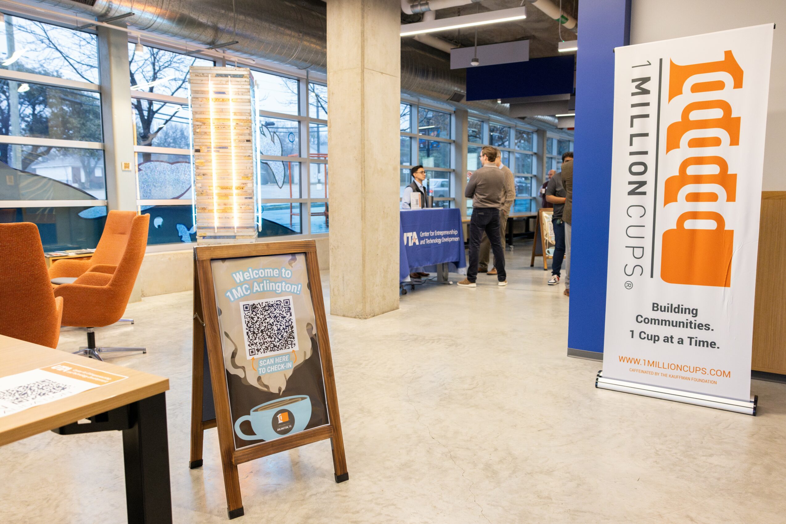 Indoor event space with people, banners, and seating, featuring large windows and modern decor.