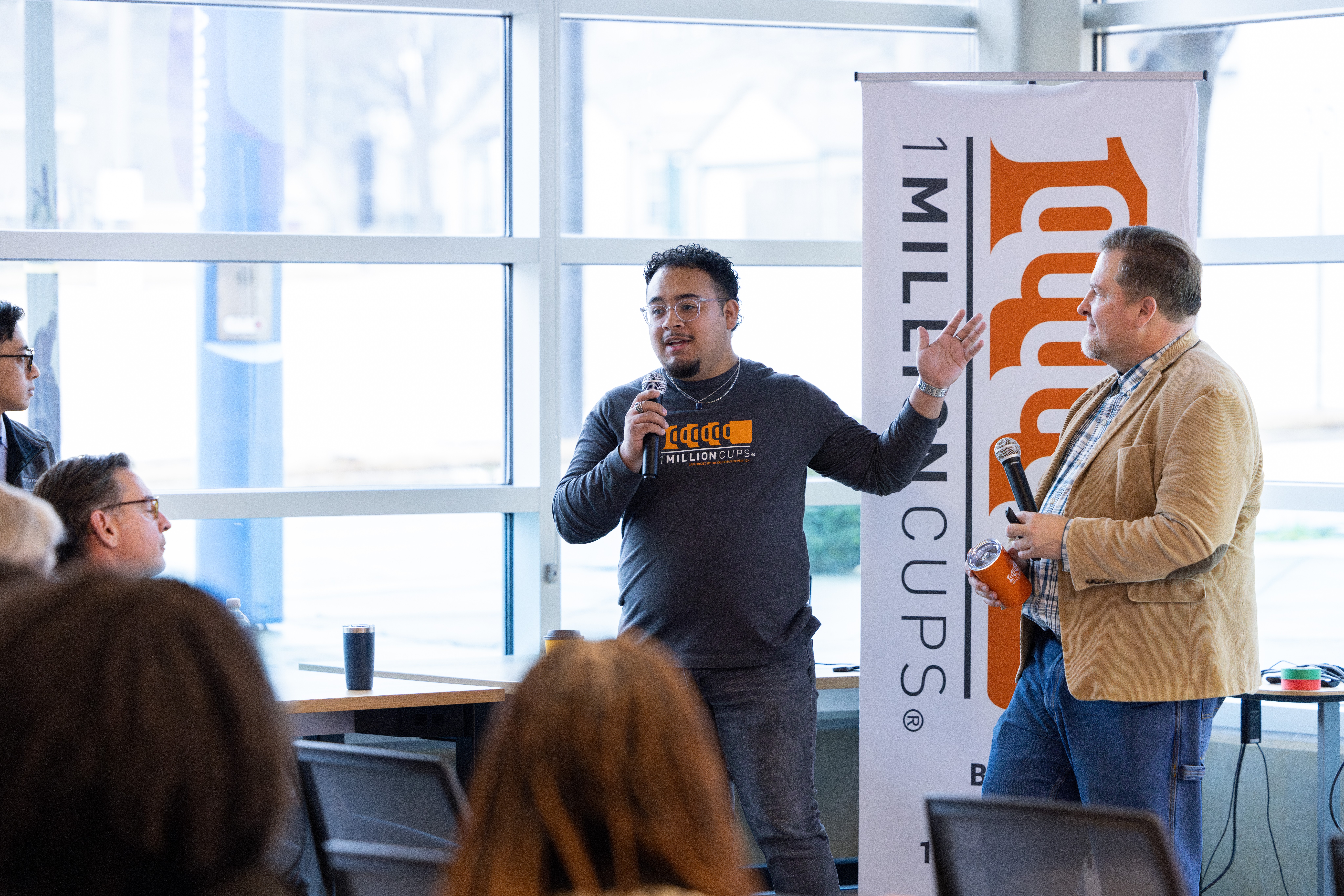 Man speaking into microphone, another man holding a paper, audience seated, large windows in background, event banner nearby.