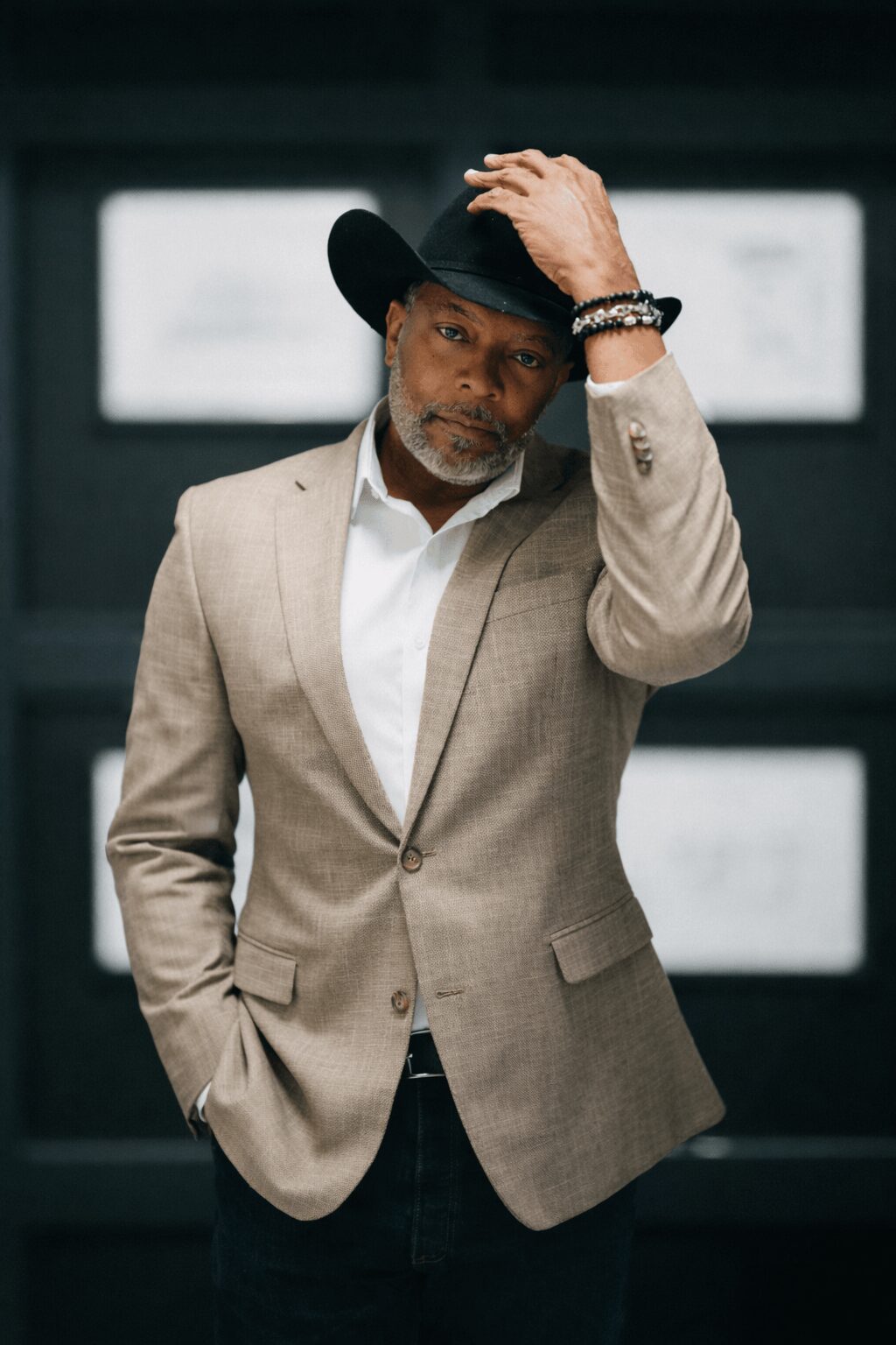 Man in beige blazer and white shirt, wearing a black hat, standing indoors with hand on hat, looking at camera.