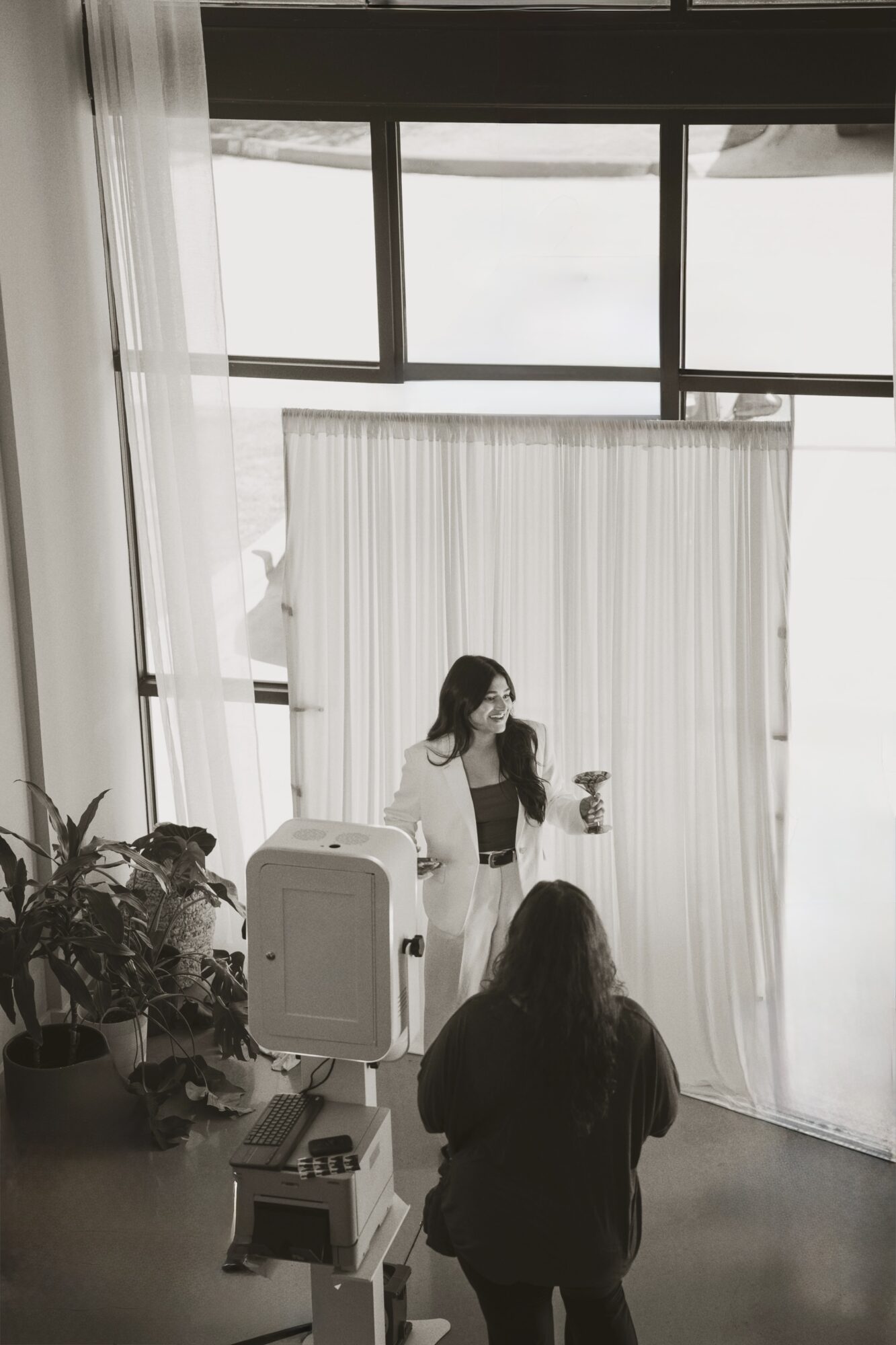 Woman standing on stage holding a microphone, audience member in foreground, large window with curtains behind, indoor setting.