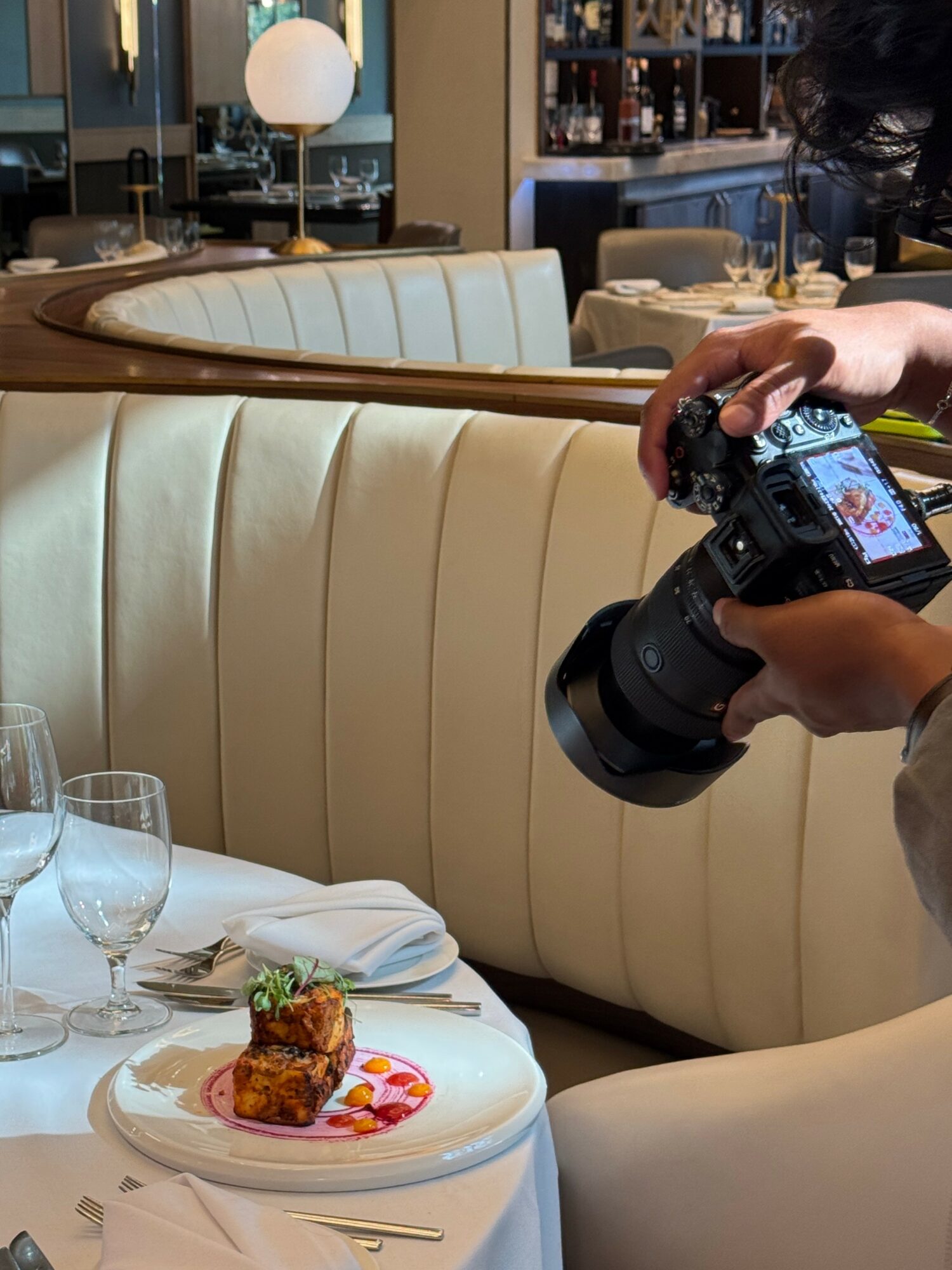 Person taking a photo of a plated dish at a restaurant table with glasses and napkins, booth seating in background.