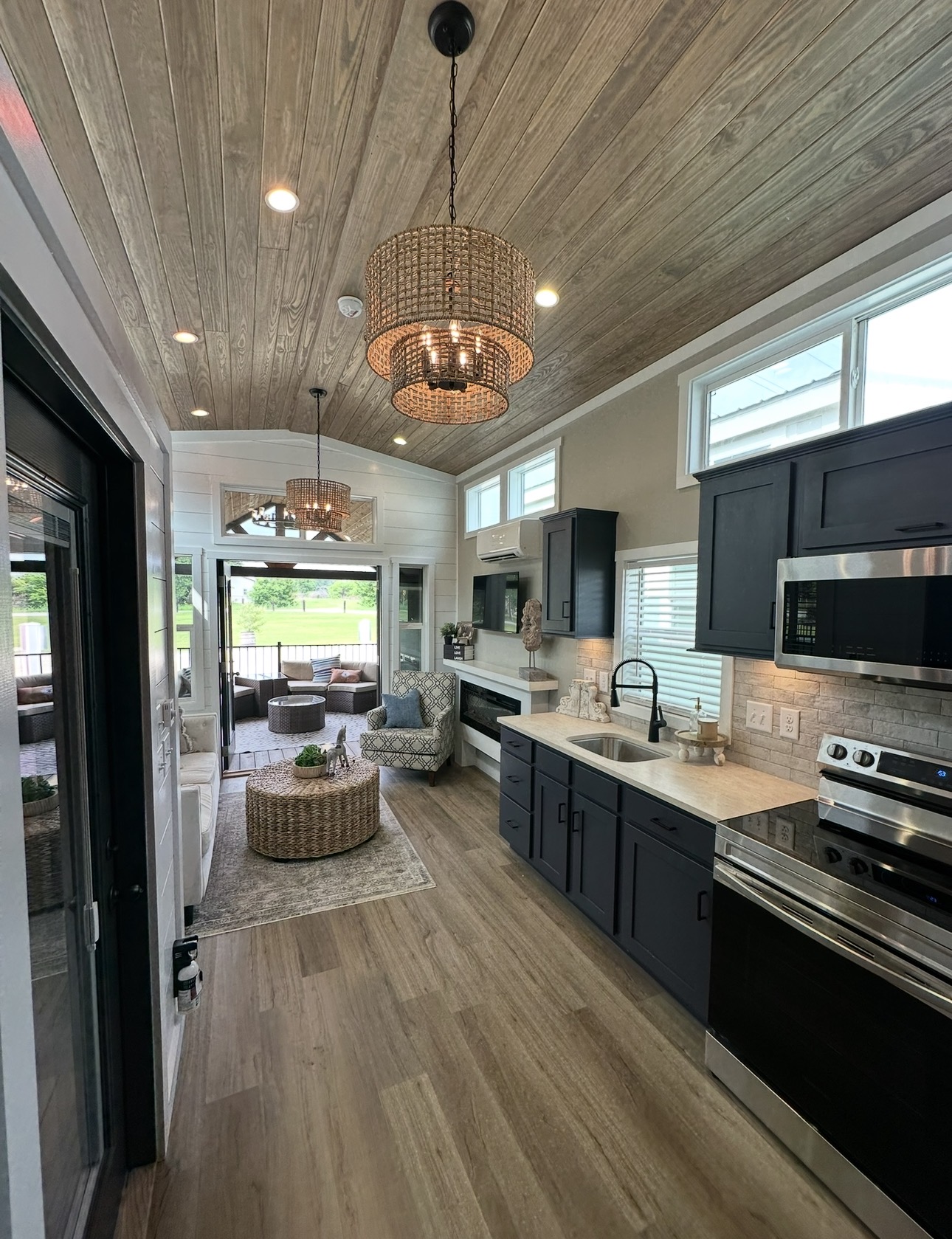 Kitchen with dark cabinets, light countertops, and a view into a living area with a rug and seating, ceiling with wood paneling, and large windows.