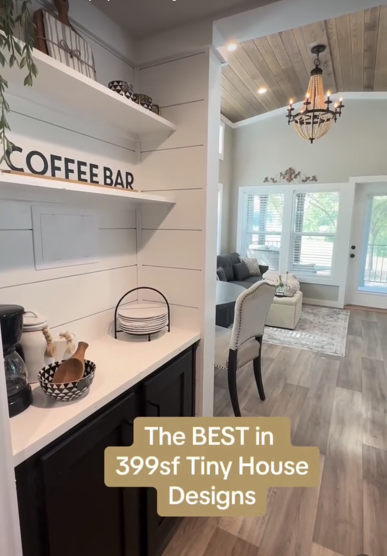Interior view of a tiny house with a coffee bar, dining area, and living room with large windows and wooden floors.