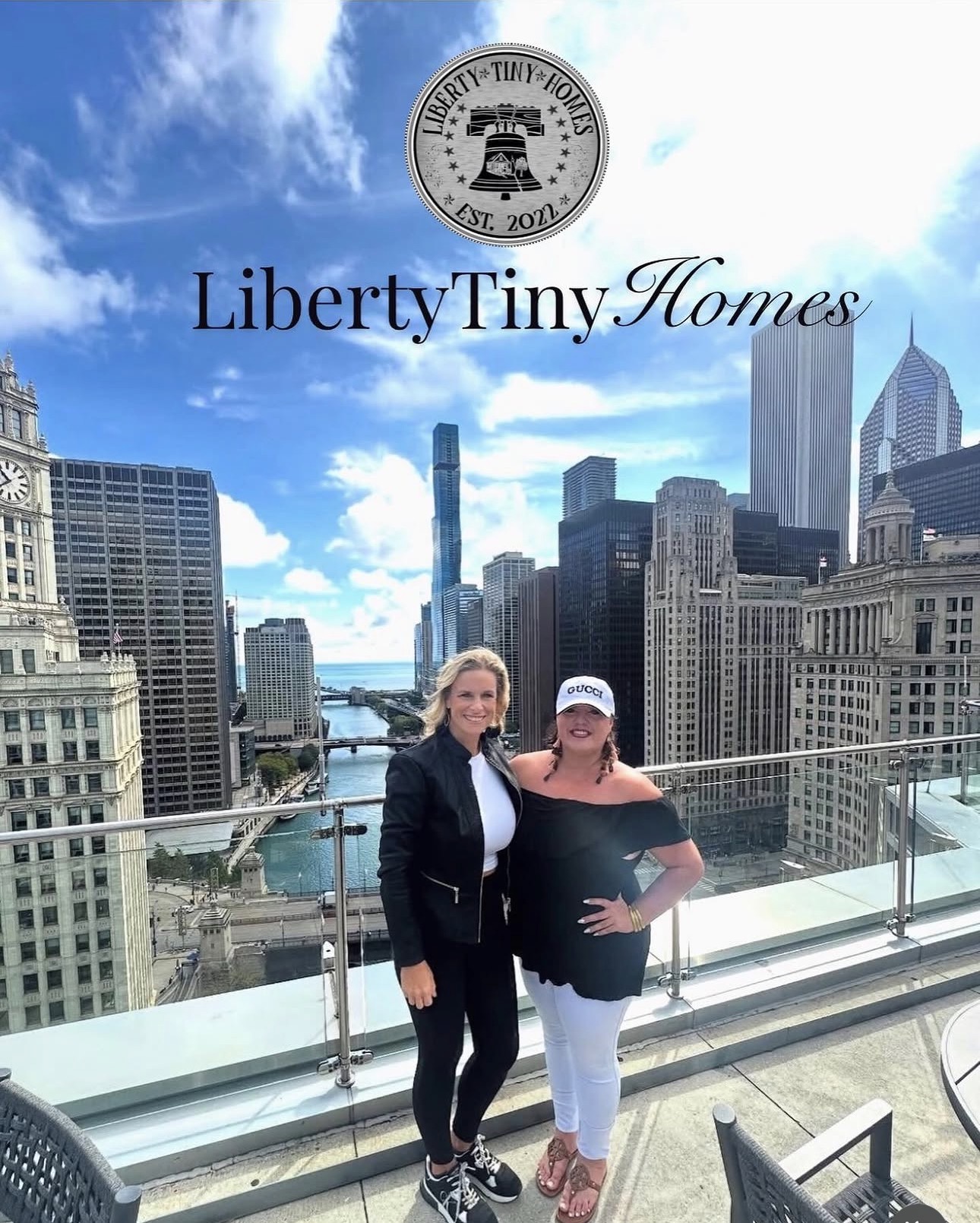 Two women stand on a rooftop with a city skyline and river behind them, smiling at the camera.