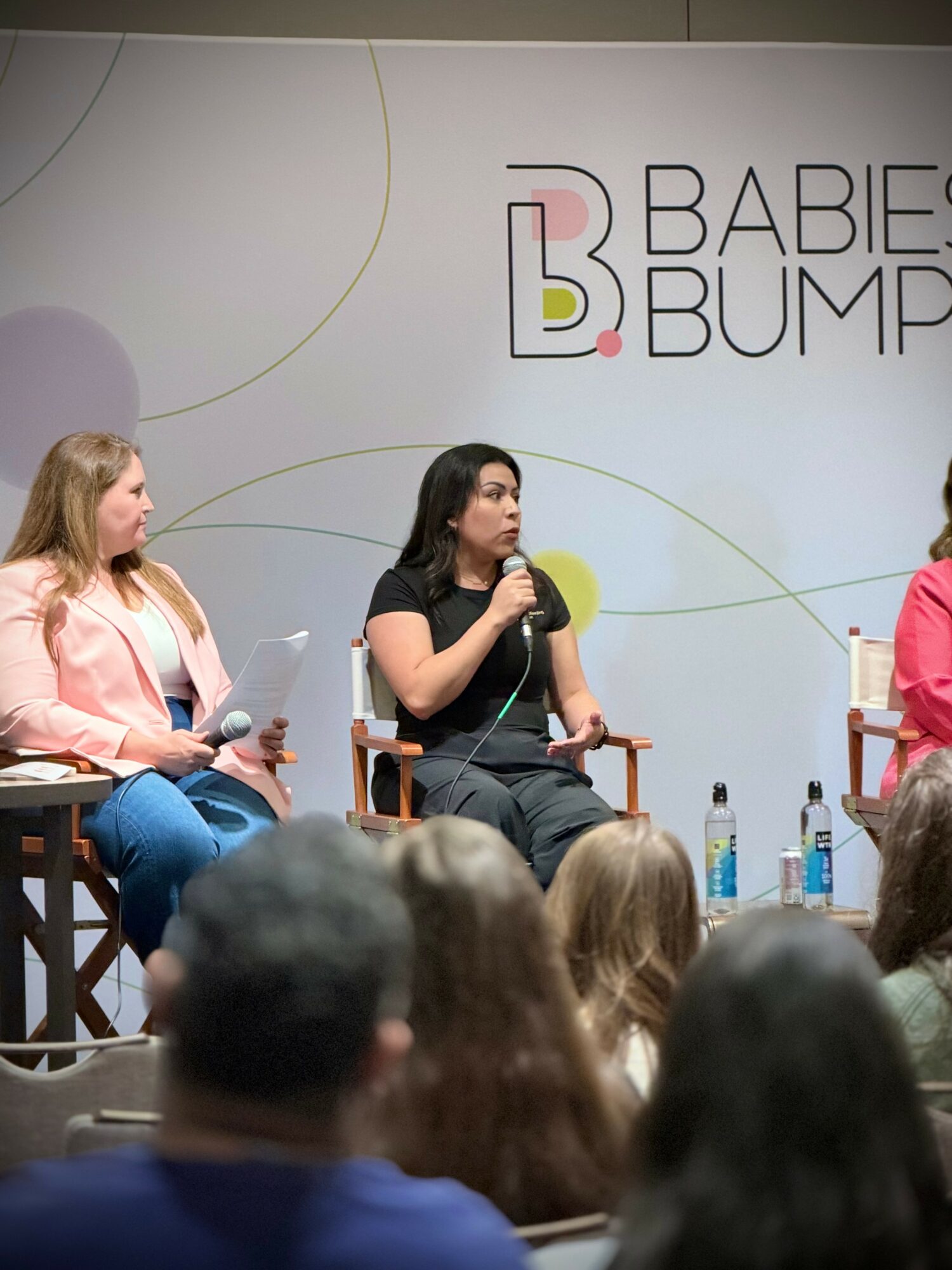Three women sit on stage in front of a panel, speaking into microphones, with an audience in front. A sign reads 'BABIES BUMP'.