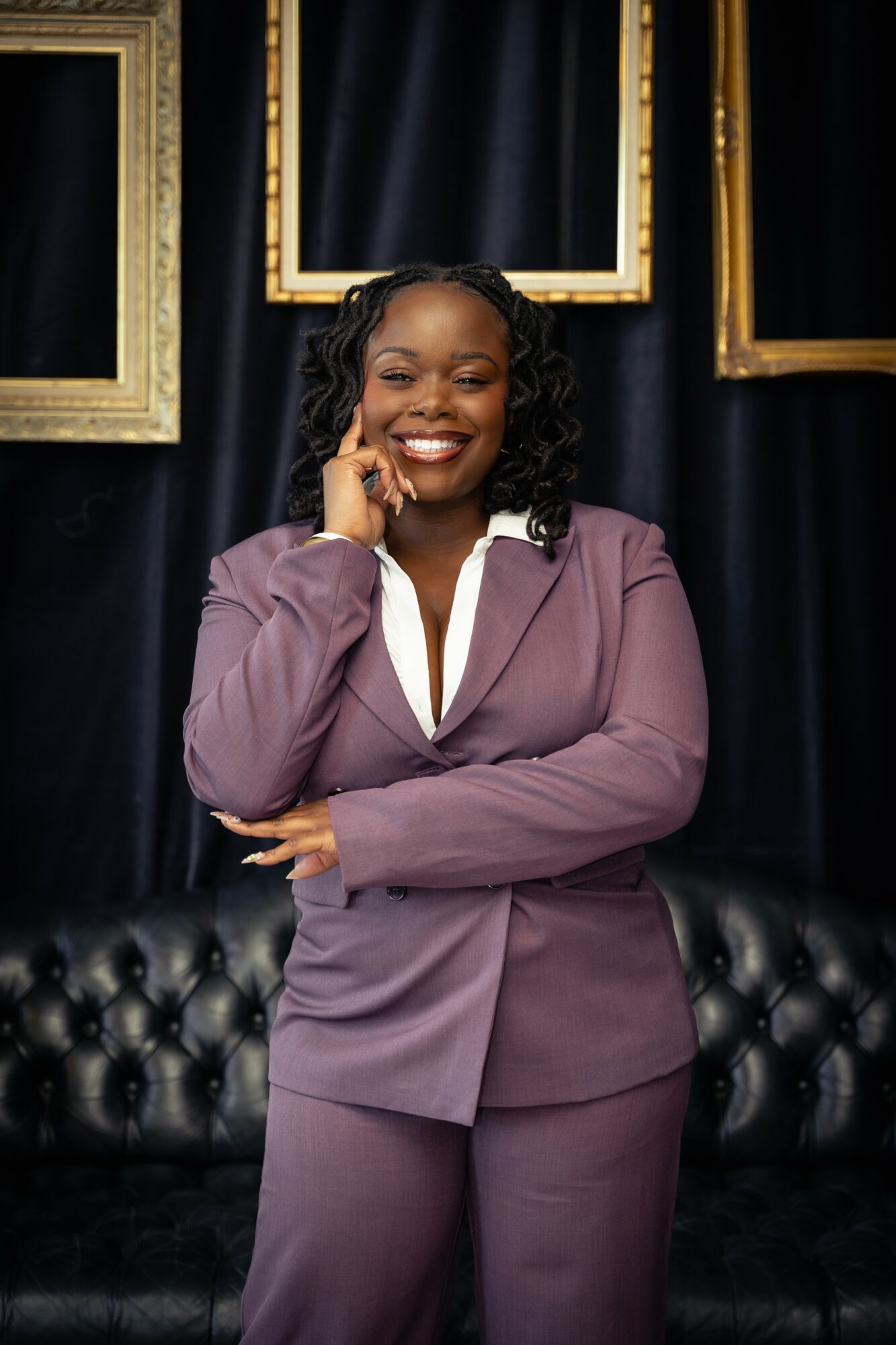 Smiling woman in purple suit standing in front of black curtain with gold frames, arms crossed, holding phone to her ear.