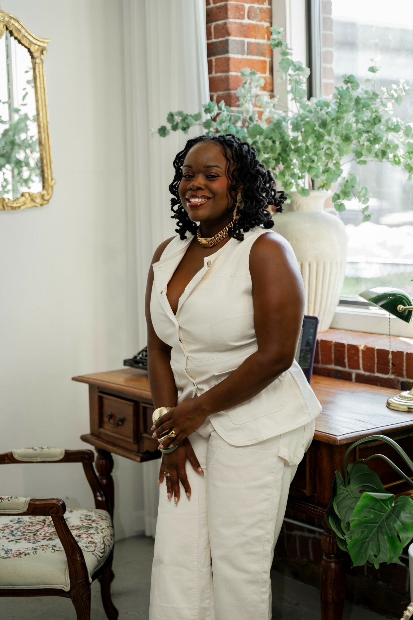 Woman in white sleeveless suit smiling, standing indoors near a window with plants and brick wall, holding hands in front.