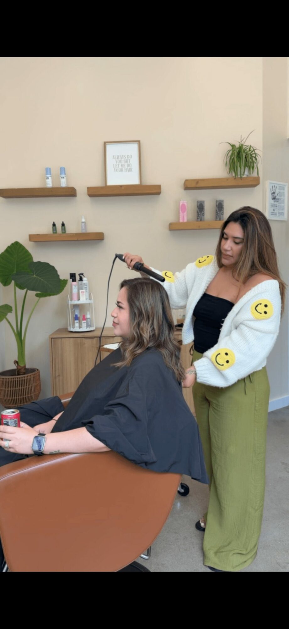 A woman is styling a young girl's hair in a salon, with shelves, plants, and framed pictures in the background.