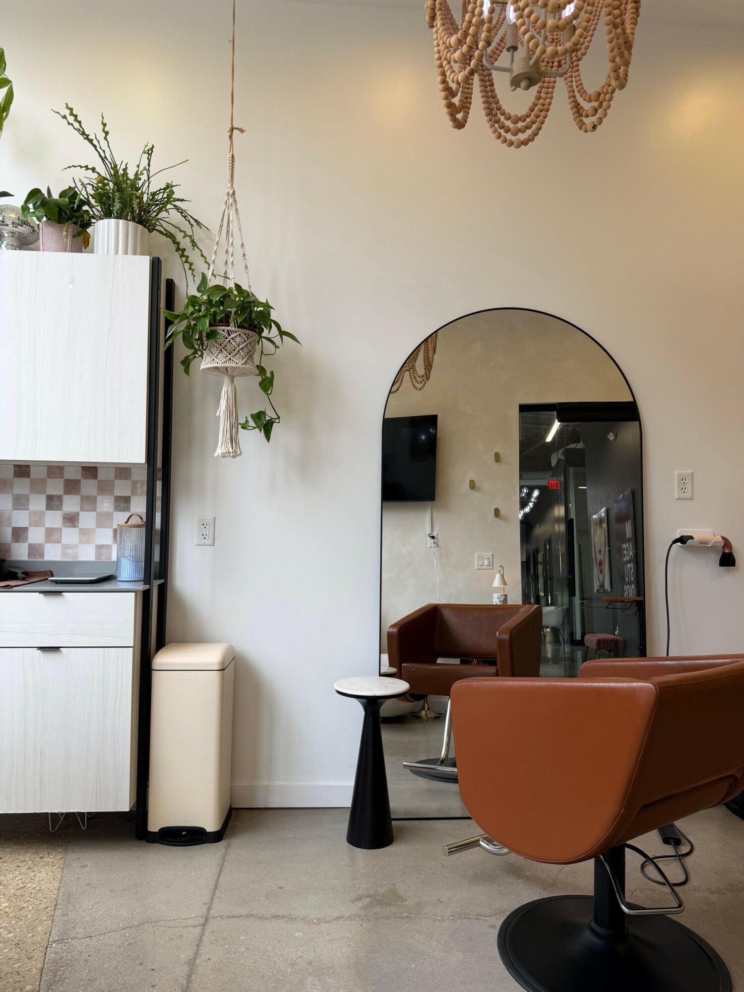 Salon with brown chair, mirror, plants, and a white cabinet, with a chandelier hanging from the ceiling.