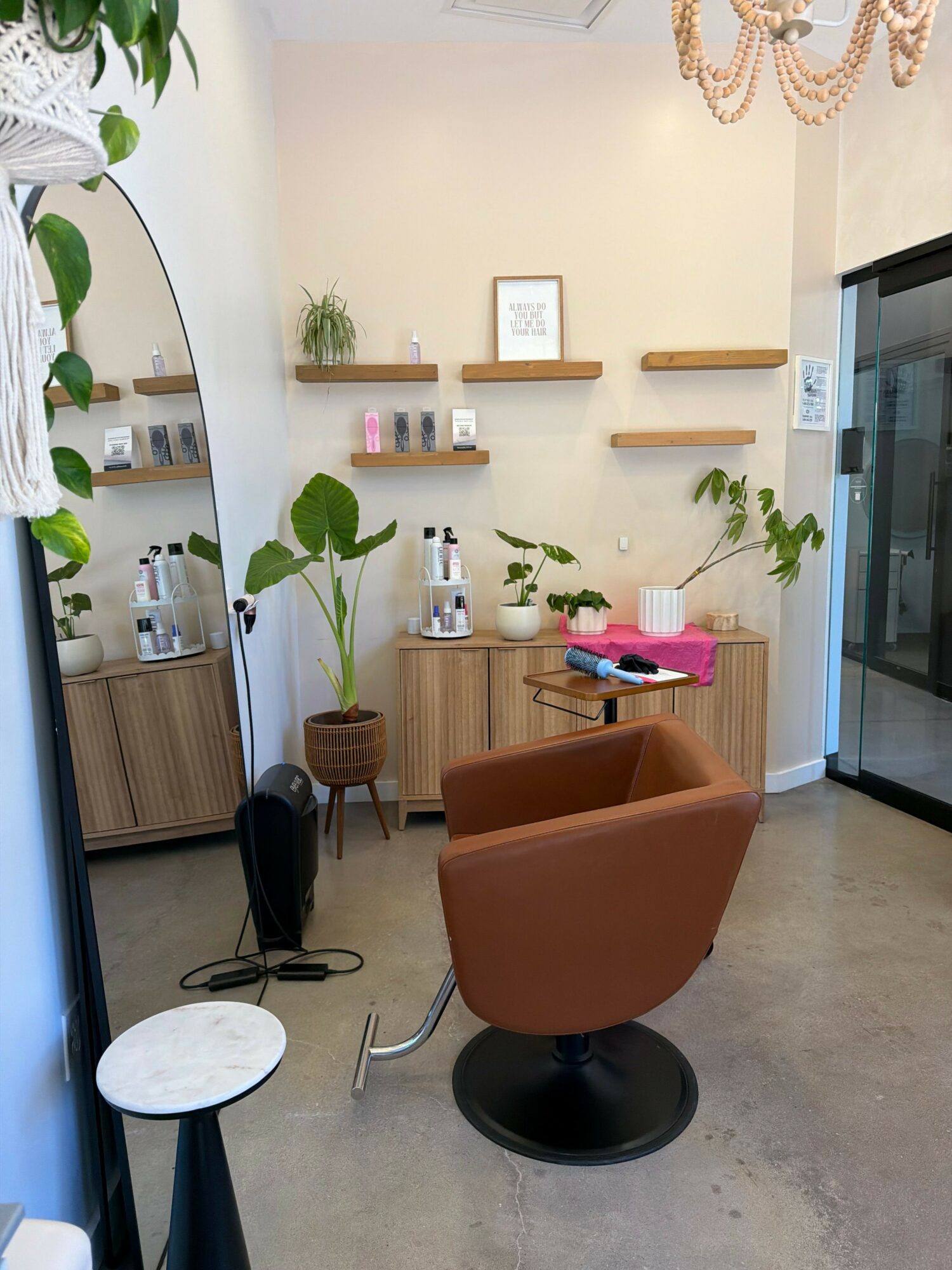 A cozy room with a brown chair, plants, shelves, and a mirror, viewed from the side. The room has a light-colored wall and floor.