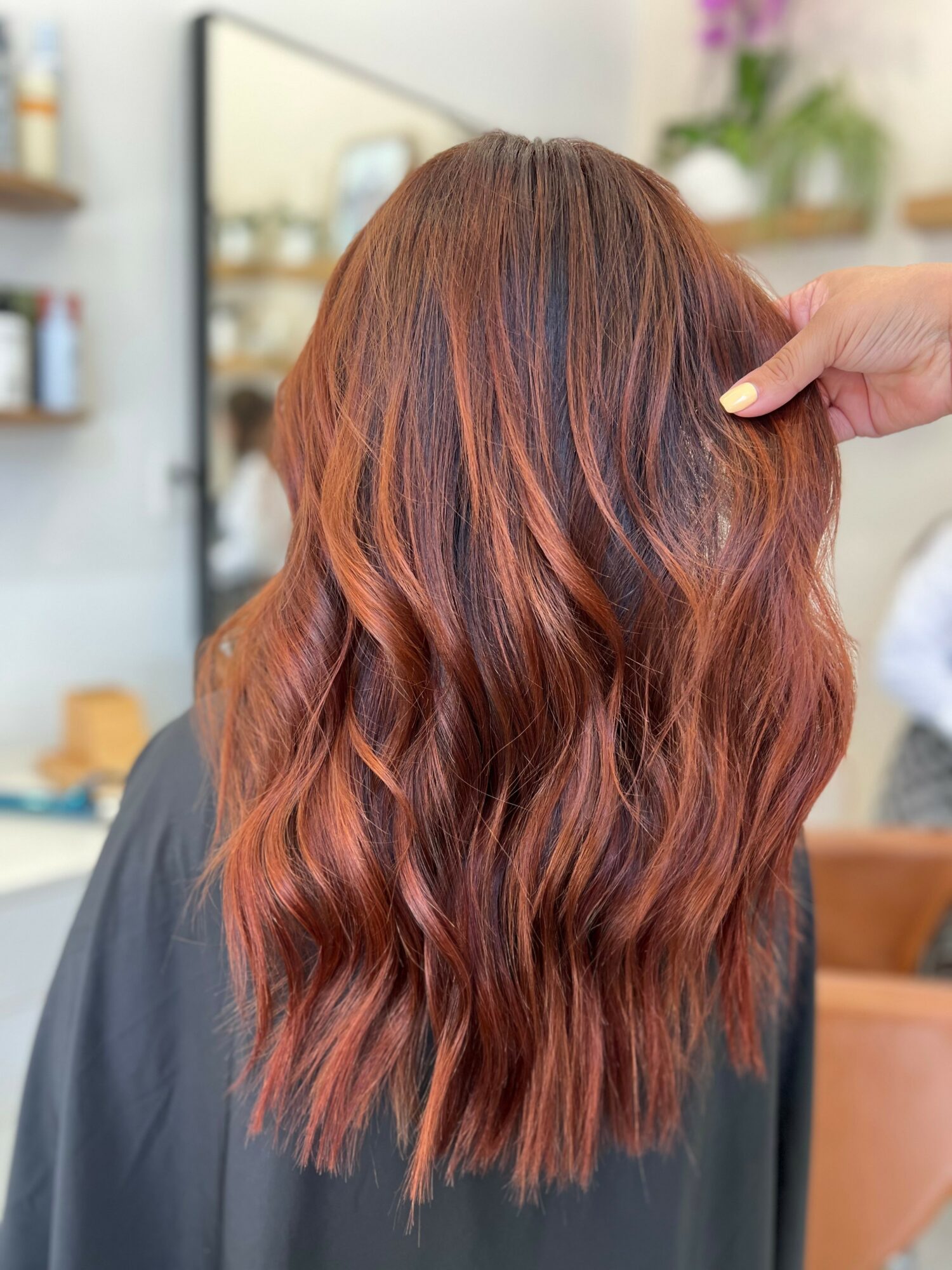 Back of a person's head with long, wavy, reddish-brown hair in a salon setting.