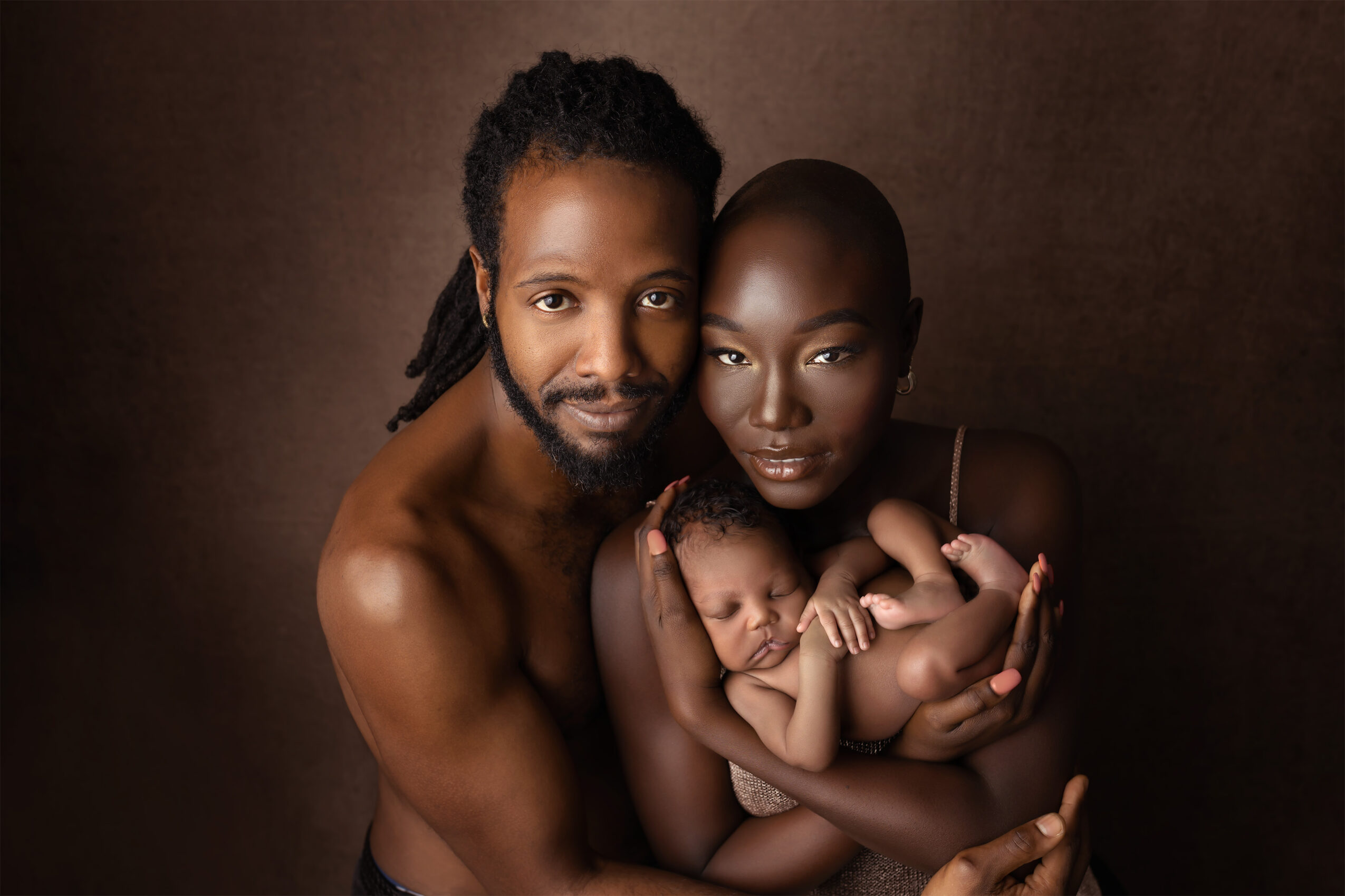 A family of three with dark skin, a man, woman, and baby, close together against a dark background.