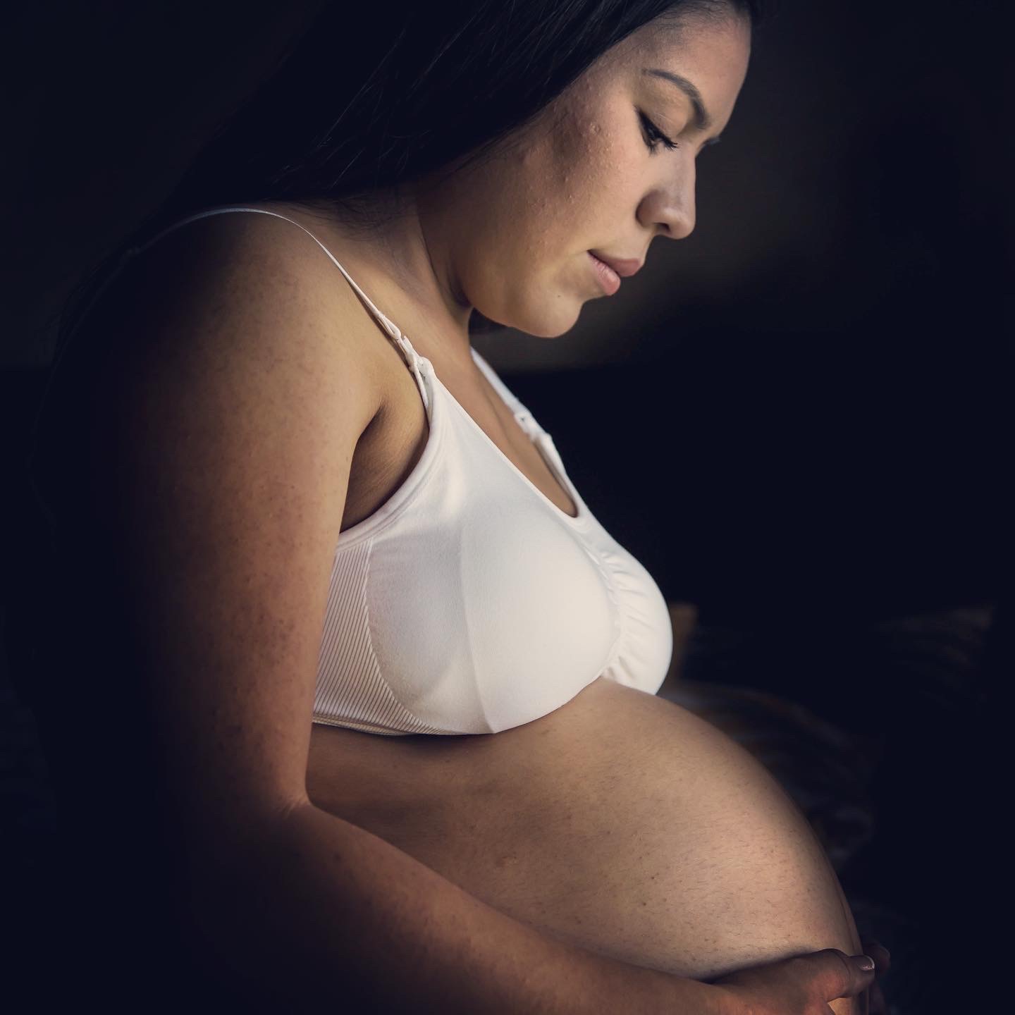 A pregnant woman in a white top looks down at her belly against a dark background.