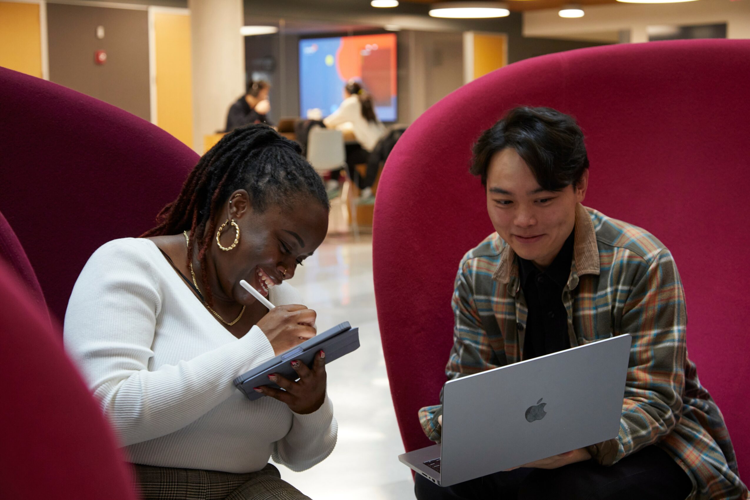 Two people sitting in large red chairs, one writing in a notebook and the other using a laptop, in a modern indoor space.