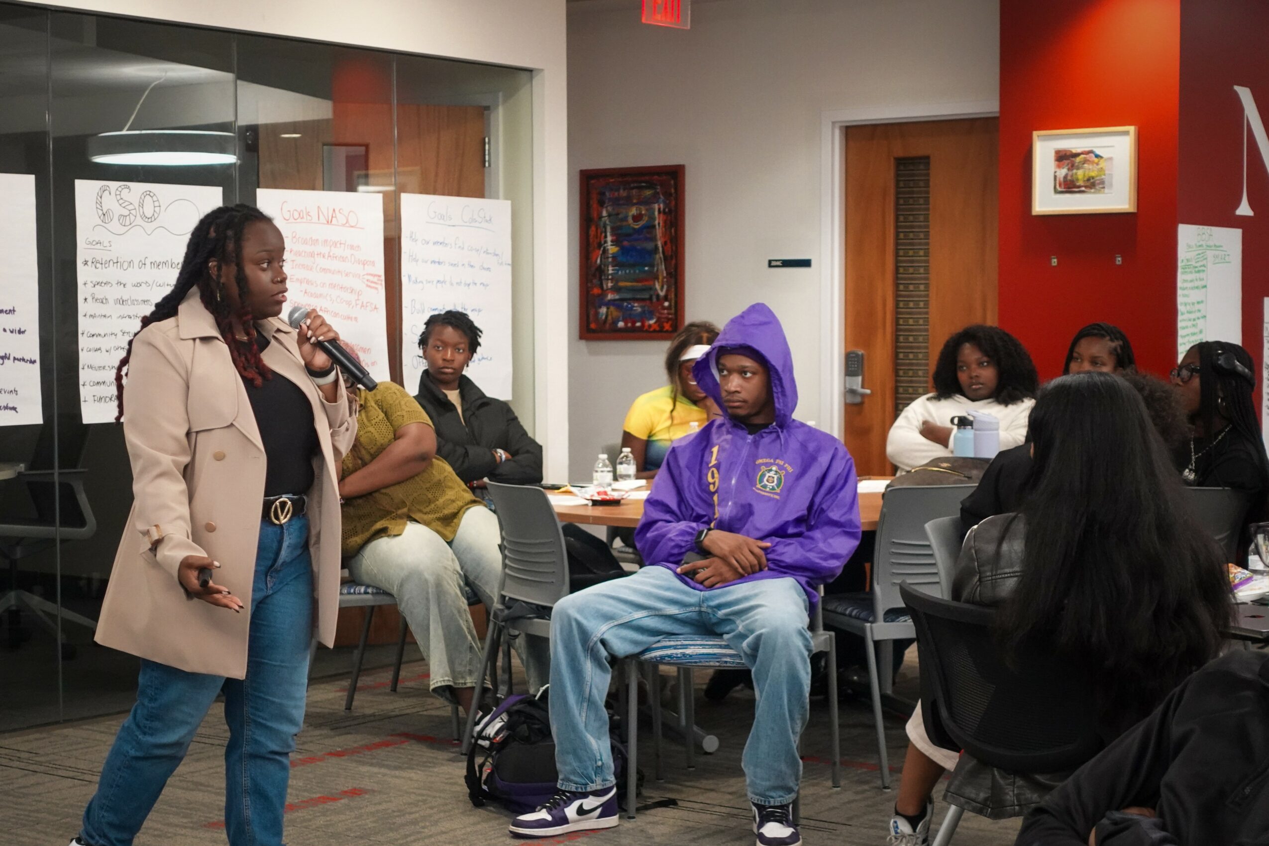 Woman speaking into microphone in a room with seated audience members, some taking notes, colorful walls, and artwork.