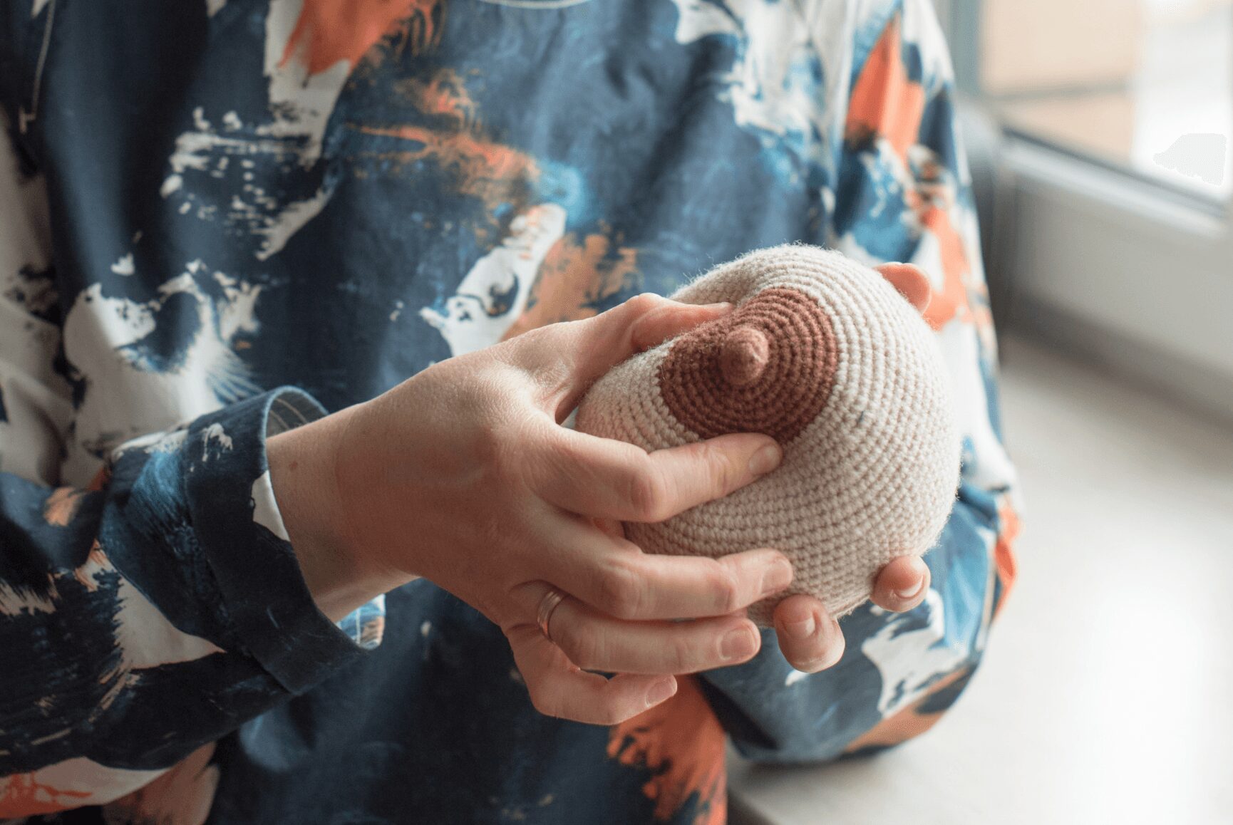 Person holding a knitted heart-shaped object with both hands, wearing a colorful patterned shirt, near a window.
