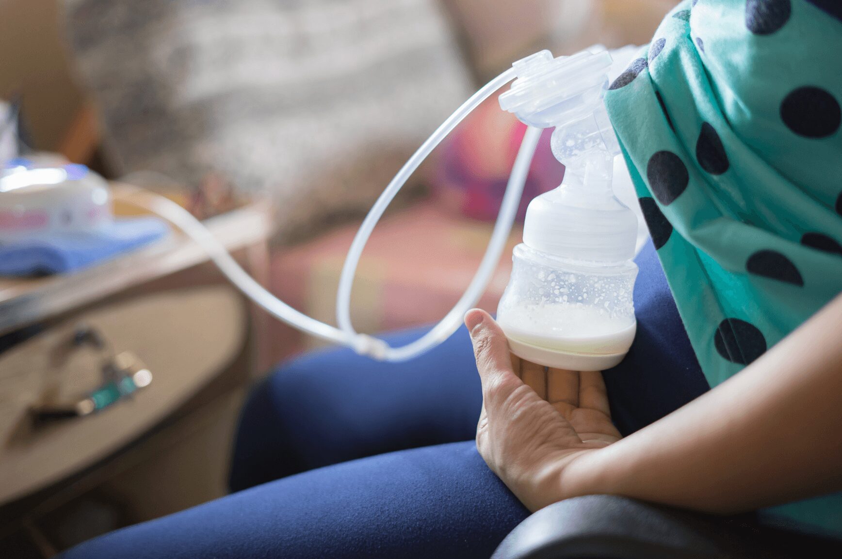 Person holding a breast pump with milk collection bottle, wearing a teal polka dot shirt, sitting indoors.