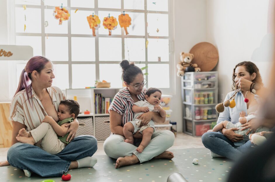 Group of women and children sitting on the floor in a bright room with a window and toys.